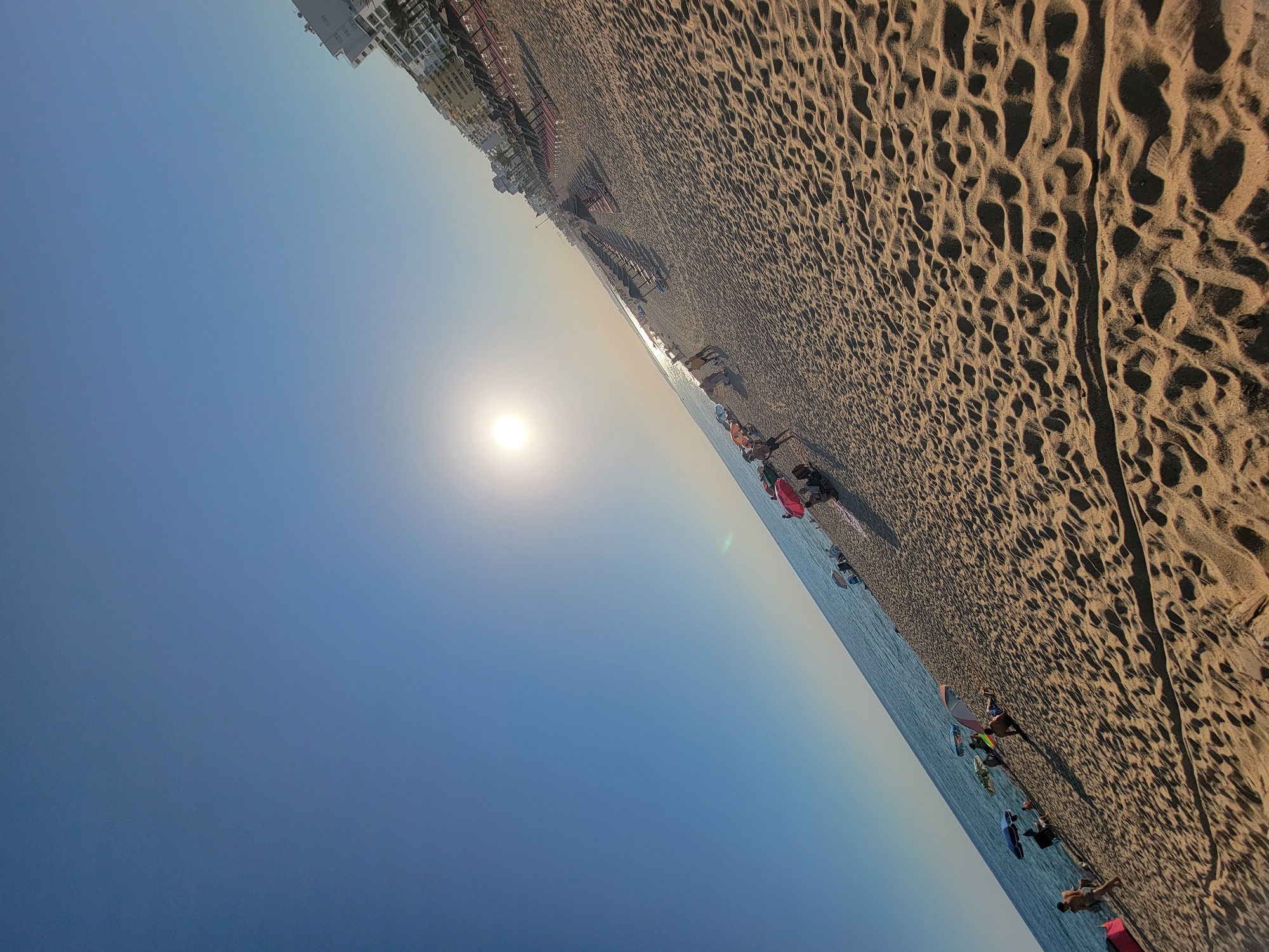 The image shows a wide shot of a sandy beach in Quarteira, Portugal, during the day. The sun is visible in the sky, casting a warm glow. The beach is populated with several people, some relaxing under beach umbrellas and others walking along the shoreline. In the background, there are buildings and rows of lounge chairs. The sand is textured with footprints and ripples. The scene depicts a typical day at the beach with recreational activities.