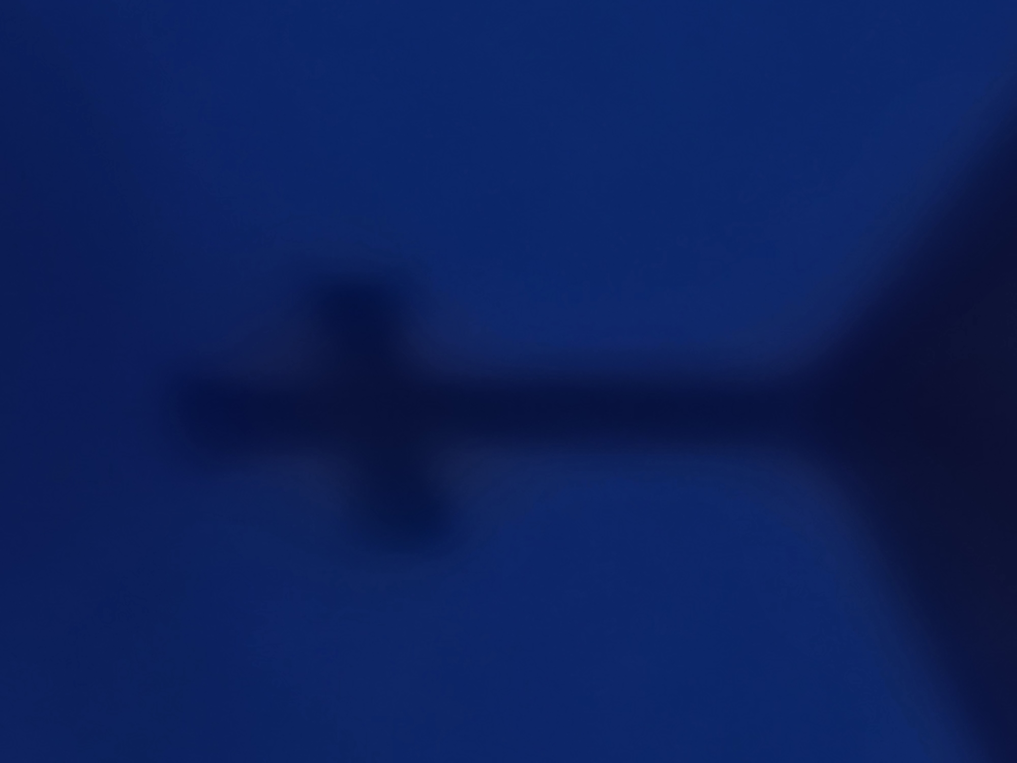 The image shows a blurred silhouette of a cross casting a shadow against a dark blue background. The shadow appears to be cast on an angled surface, possibly the roof of a building. The overall impression is of a dimly lit, possibly evening or nighttime scene. The location in Gura Top, Nigeria, is not discernible from the image itself.