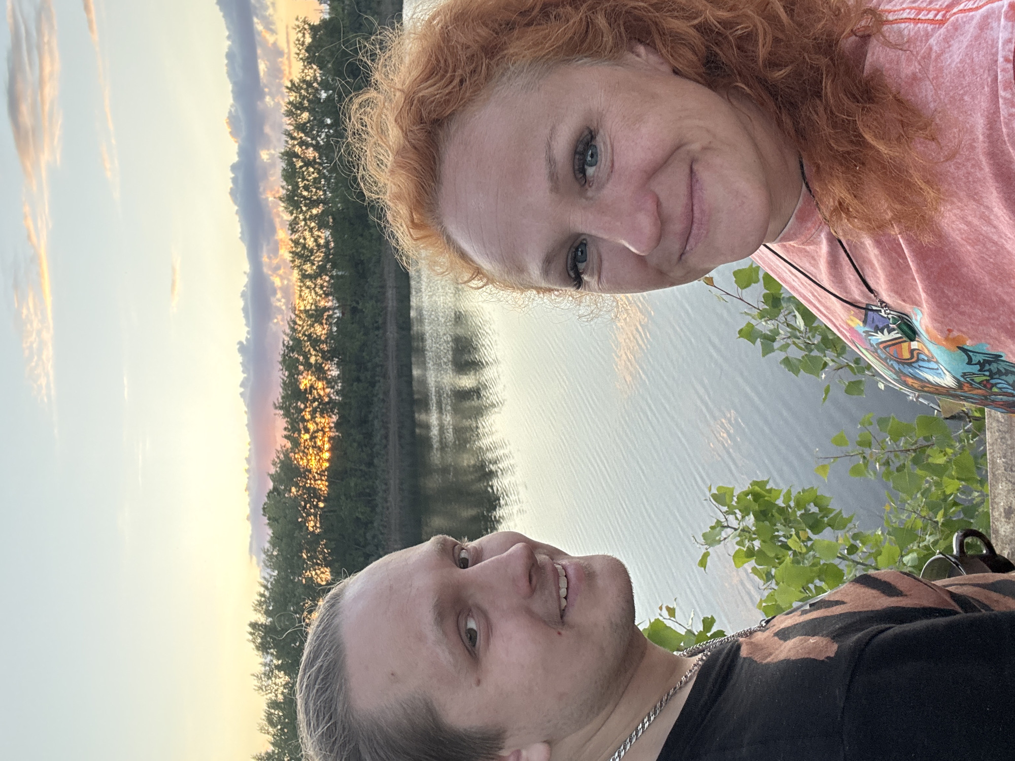 This image is a selfie-style portrait featuring two individuals, an adult woman and an adult man, set against an outdoor backdrop during sunset or dusk. The photograph is oriented vertically, but when viewed upright, the woman is positioned in the upper right quadrant of the frame, and the man is in the lower left.
The woman, with distinct curly auburn hair, looks directly towards the camera with a gentle, contented smile. She has blue eyes and is wearing a light-colored, possibly pinkish-orange, t-shirt that features a colorful graphic design on the chest. A dark necklace with a prominent green pendant is visible around her neck.
The man, with lighter hair, is looking slightly upwards and to his right, offering a subtle smile. He is wearing a black t-shirt with a brown or orange pattern, and a silver chain necklace is visible around his neck.
Behind the two people, a wide body of water, likely a river or lake, reflects the soft, warm light of the setting sun. A dense line of trees forms the distant bank, silhouetted against the sky, which displays hues of orange, pink, and pale blue, characteristic of either sunrise or sunset. Some green foliage is also visible in the immediate foreground on the right.
The scene conveys a peaceful and relaxed atmosphere, suggesting a shared moment outdoors. This tranquil setting is located in Chaikovsky, Russia. There is no discernible text visible within the image.