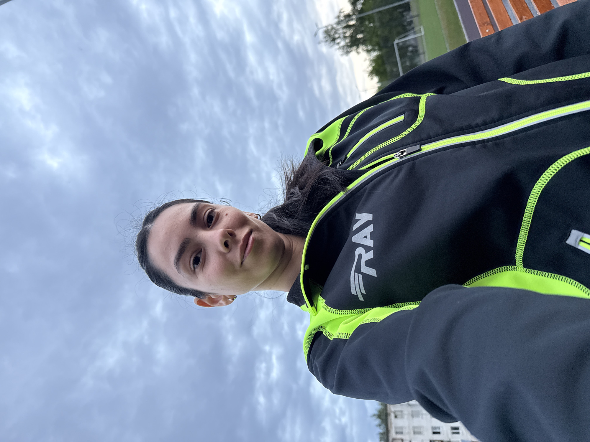 A woman is seen in a low-angle selfie. She is wearing a black jacket with neon green accents and the word "FRAY" printed on it. The background shows a cloudy sky, and there is a grassy field with a soccer goalpost and a bench visible in the distance. A multi-story building is also present in the background. Given the information provided, the location is Chaikovsky, Russia.
