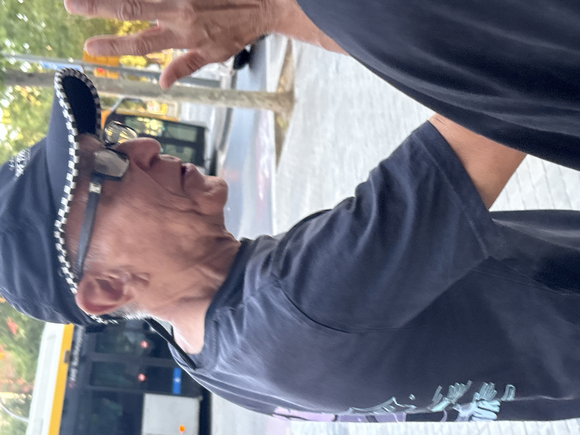 An outdoor, eye-level, tilted shot captures an older man from the chest up in what appears to be an urban street setting in Barcelona, Spain. He wears a dark blue or black t-shirt with faint, light-colored graphics resembling stylized hands on the lower left side. A matching dark cap with a distinctive black and white checkered pattern along its brim sits on his head. Dark-framed glasses with reflective lenses are perched on his nose. His mouth is open, suggesting he is speaking or reacting to something, and his gaze is directed upwards and to the right of the frame. In the upper right corner, another person's open hand is visible, possibly mid-gesture or in interaction with the main subject. The background is softly blurred but reveals green foliage from trees, a yellow stripe indicative of a public bus, and general street elements, all illuminated by bright daylight. No discernible text is visible anywhere in the image.