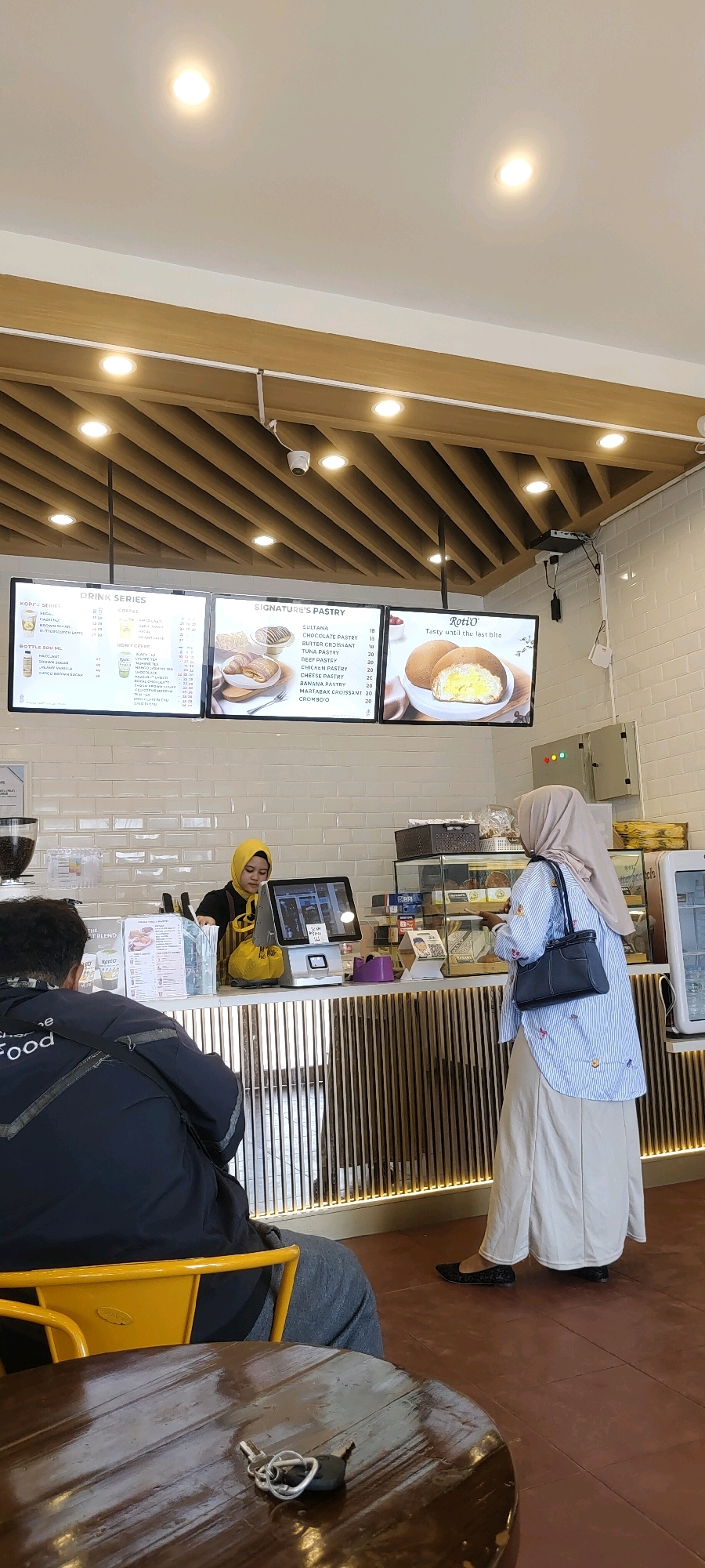 The image captures an indoor scene, likely a cafe or bakery, with two women and one man present. The environment appears to be clean and modern, with white tiled walls and a wooden slatted ceiling. The overall lighting is bright, with several recessed ceiling lights illuminating the space.
In the center of the image, a female employee wearing a yellow hijab and apron stands behind a counter. She is interacting with a customer, a woman wearing a light brown hijab and a blue and white striped shirt with colorful embroidered butterflies, and a long beige skirt. The customer is facing away from the camera, holding a black handbag. Both women are standing in front of the counter, which has a touch screen ordering system.
To the left of the counter, a man is seated in a yellow chair, facing away from the camera. He is wearing a dark jacket with white lettering on the back that reads "Food". He is seated at a round, dark wooden table. A set of keys lies on the table in front of him.
Behind the counter, digital menu boards are mounted on the wall. One board displays "DRINK SERIES", another "SIGNATURE'S PASTRY", and a third "Roti'O". The pastry board lists items like "SULTANA", "CHOCOLATE PASTRY", and "BANANA PASTRY" with prices. The "Roti'O" board features an image of a pastry cut in half with filling.
On the right side of the counter, a glass display case contains various baked goods. There are also electrical boxes mounted on the wall to the right of the display case.
The scene is consistent with a commercial establishment in Banjarbaru, Indonesia, likely a popular cafe or bakery due to the menu displays and the type of food items advertised. The overall atmosphere suggests a typical customer interaction at a food service establishment.