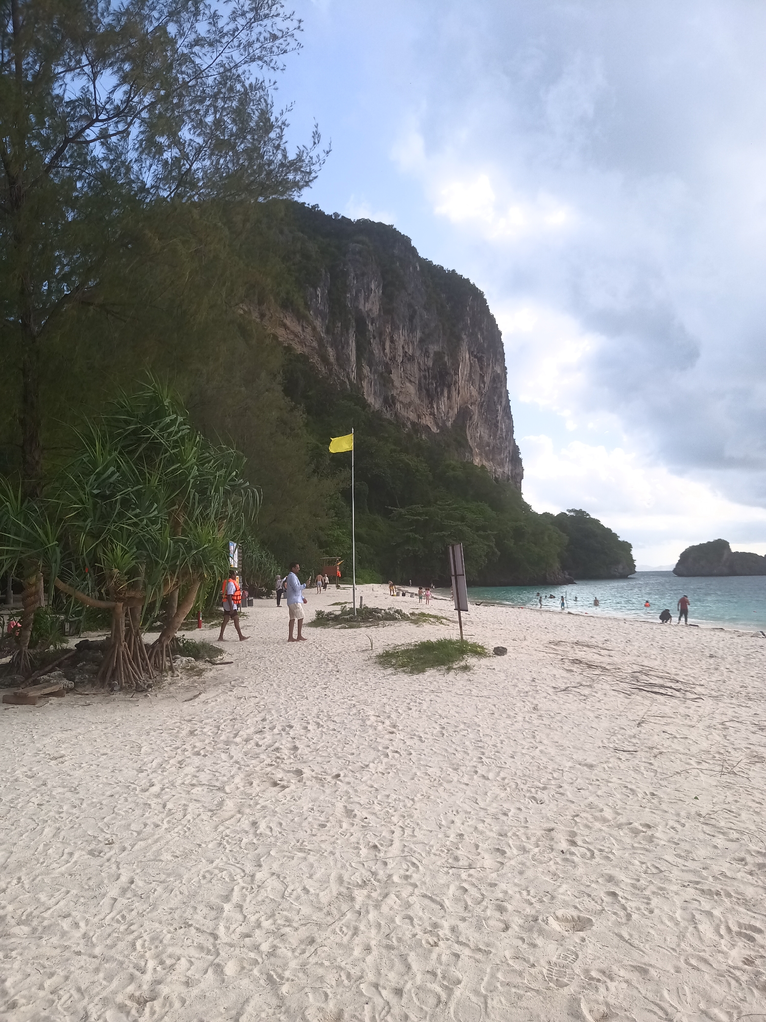 The image depicts a beach scene in Ban Ao Nang, Thailand. The beach has light-colored sand with sparse vegetation. In the background, a large limestone cliff rises vertically, covered in green foliage on the lower portion. The sky is overcast with a mix of blue sky and gray clouds. A yellow flag is planted in the sand. Several people are present, including individuals walking along the shore, some wearing life vests, and others swimming in the ocean. Debris, including twigs and small pieces of wood, litters the beach.