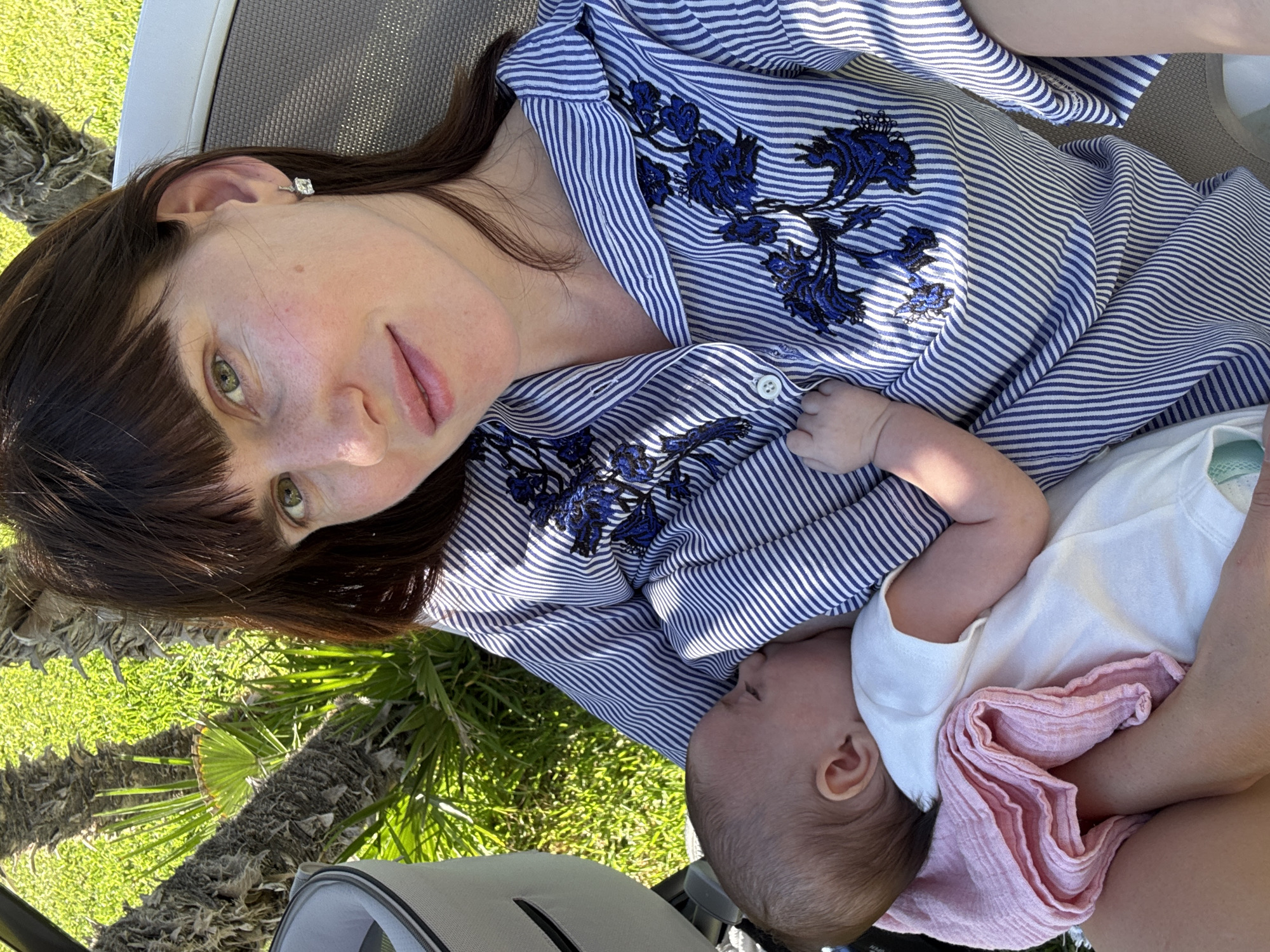 A woman with dark brown hair and green eyes, wearing a blue and white striped blouse with blue floral embroidery, is breastfeeding a baby outdoors. The woman is seated in a lounge chair, with a baby stroller to her left. The background consists of green grass and palm trees, suggesting a sunny, warm climate. The overall scene conveys a moment of intimacy and care between mother and child in a relaxed, outdoor setting, likely within a garden or park area. The city of Capaci, Italy, is known for its beautiful coastal scenery and lush vegetation, which aligns with the visual cues present in the image.