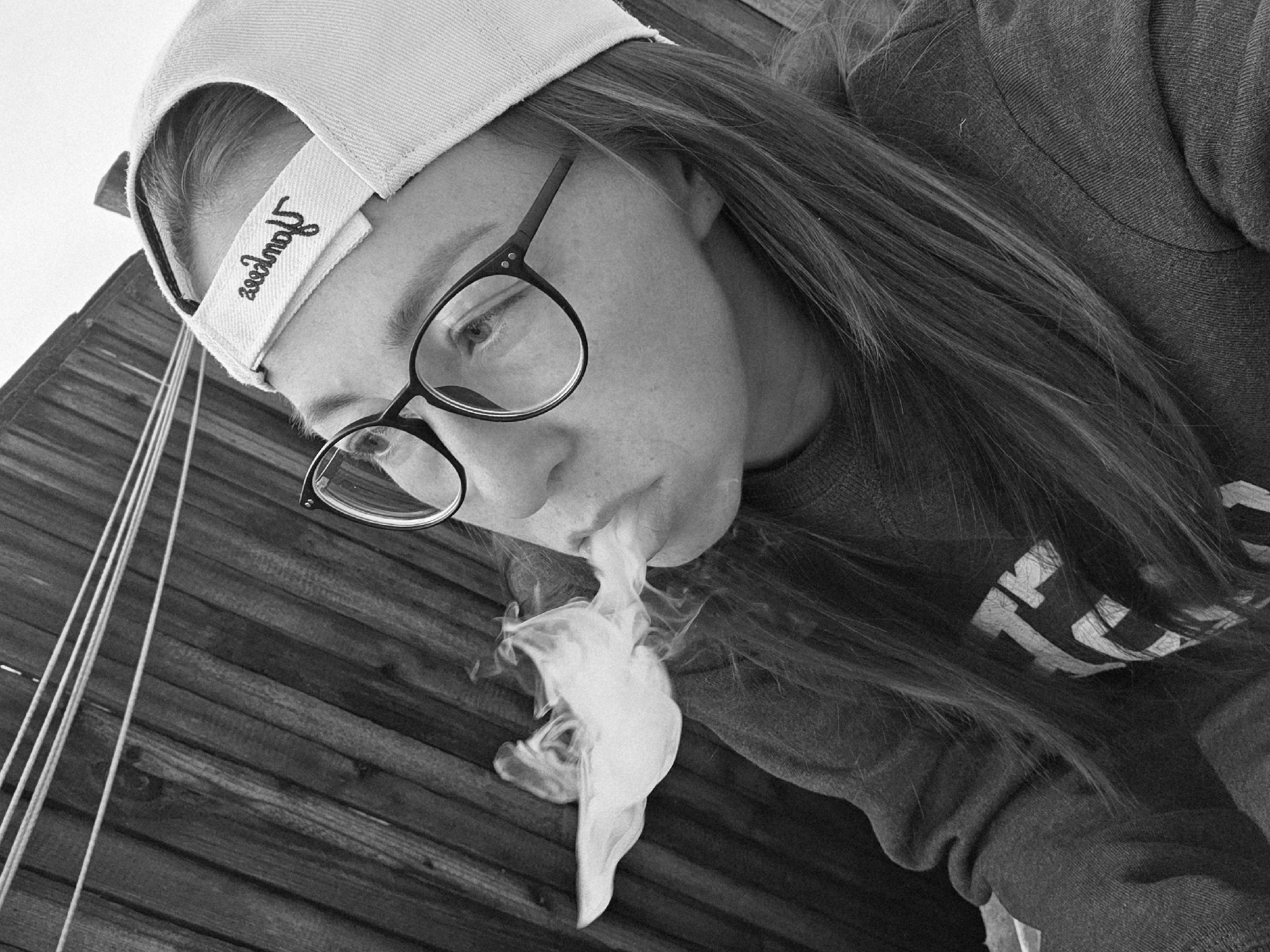 The black and white image shows a person wearing glasses and a baseball cap, exhaling smoke. They are standing in front of a wooden fence. The person is wearing a dark sweatshirt with white lettering on it. The scene appears to be outdoors. There are no discernable landmarks or indicators that would place the image in the city of Chaikovsky, Russia.