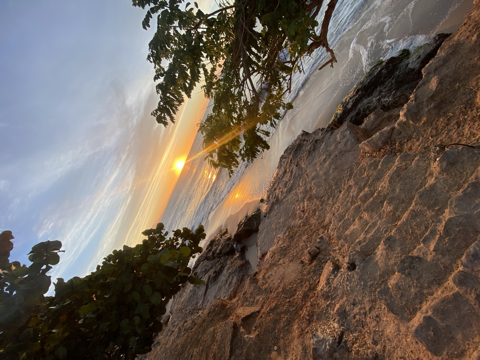 The image depicts a sunset scene at a beach in Juan Griego, Venezuela. The camera is angled upward from a rocky outcrop, showcasing its textured surface in the foreground. To the left and above, green foliage from trees or bushes frames the scene. The setting sun casts a golden hue across the ocean, with its reflection visible on the water's surface. The sky transitions from blue to orange with thin clouds. The beach itself is a narrow strip where the waves are breaking. The scene lacks any human presence or activity.
