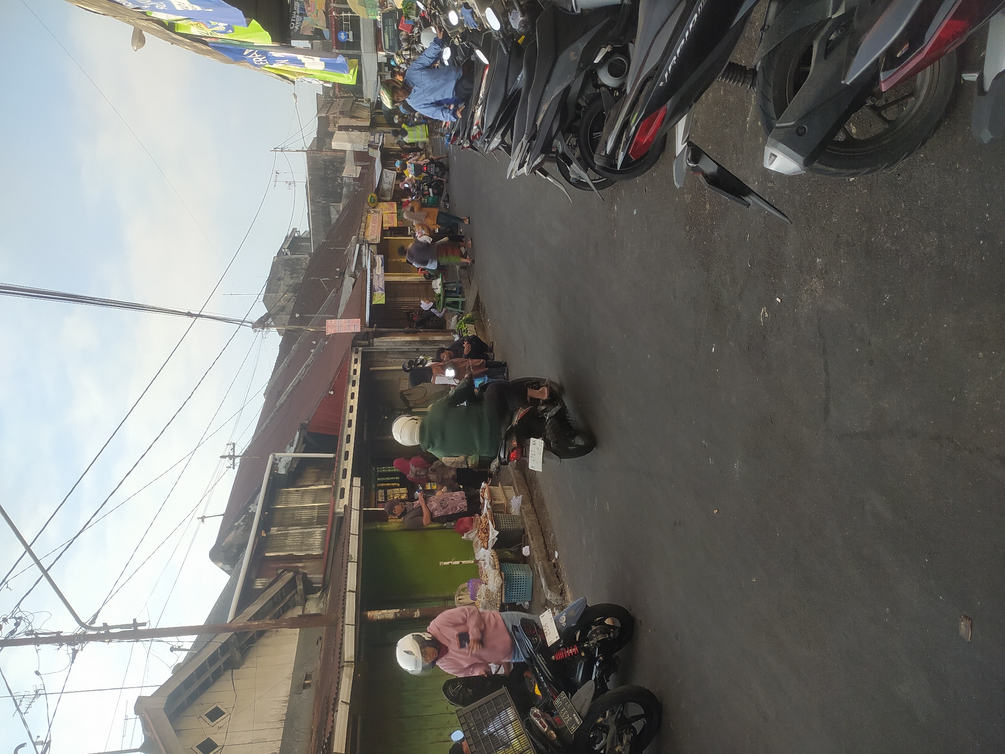 The image depicts a street scene in Salatiga, Indonesia, likely a market or commercial area. The foreground is dominated by parked motorcycles, mostly black and modern models, filling the right side of the asphalt road. The road itself is a dark grey, appearing slightly worn and uneven with some debris scattered on it.
On the left side of the road, there are several individuals. One person in a pink hoodie and white helmet is standing next to a motorcycle, holding a phone and a plastic crate. Further down, a person in a green jacket and white helmet is riding a motorcycle away from the camera. To the left of this rider are stalls or shops with various goods displayed, including what appear to be dried foods and snacks. People can be seen interacting at these stalls. The buildings lining the street are modest, with corrugated metal roofs and some signs of age.
In the background, the street extends with more buildings and people, suggesting a bustling, open-air market environment. Overhead, a complex network of electrical wires and poles crisscrosses the sky, typical of urban infrastructure in Indonesia. The sky is overcast with a pale blue hue. Banners with Indonesian text hang from some of the buildings on the right side of the street, further indicating the commercial nature of the location. The overall impression is a lively, everyday street scene in an Indonesian city.