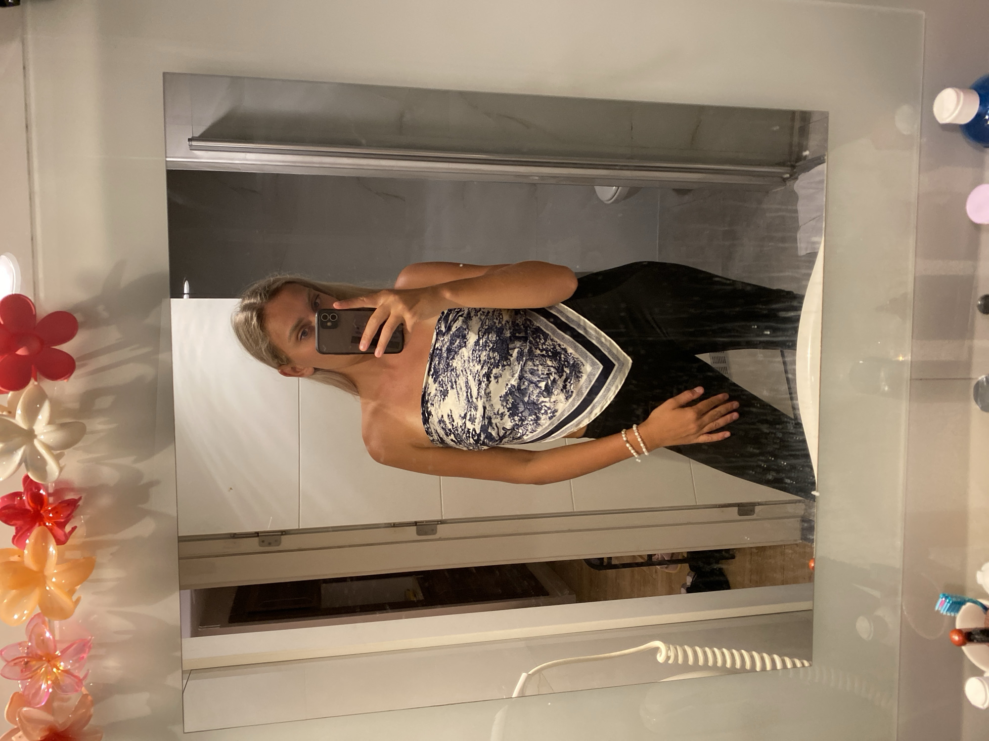 A young woman with blonde hair takes a selfie in a bathroom mirror. She is wearing a strapless top that resembles a silk scarf with a blue and white pattern, and black pants. She has a pearl bracelet on her left wrist. The mirror reflects a shower stall with a glass door and a toilet in the background. The bathroom appears to be part of a hotel or guesthouse, with decorative flower lights above the mirror. The scene suggests a personal moment captured in a private space, likely during a trip to Aksu, Turkey, given the implied context of the location.