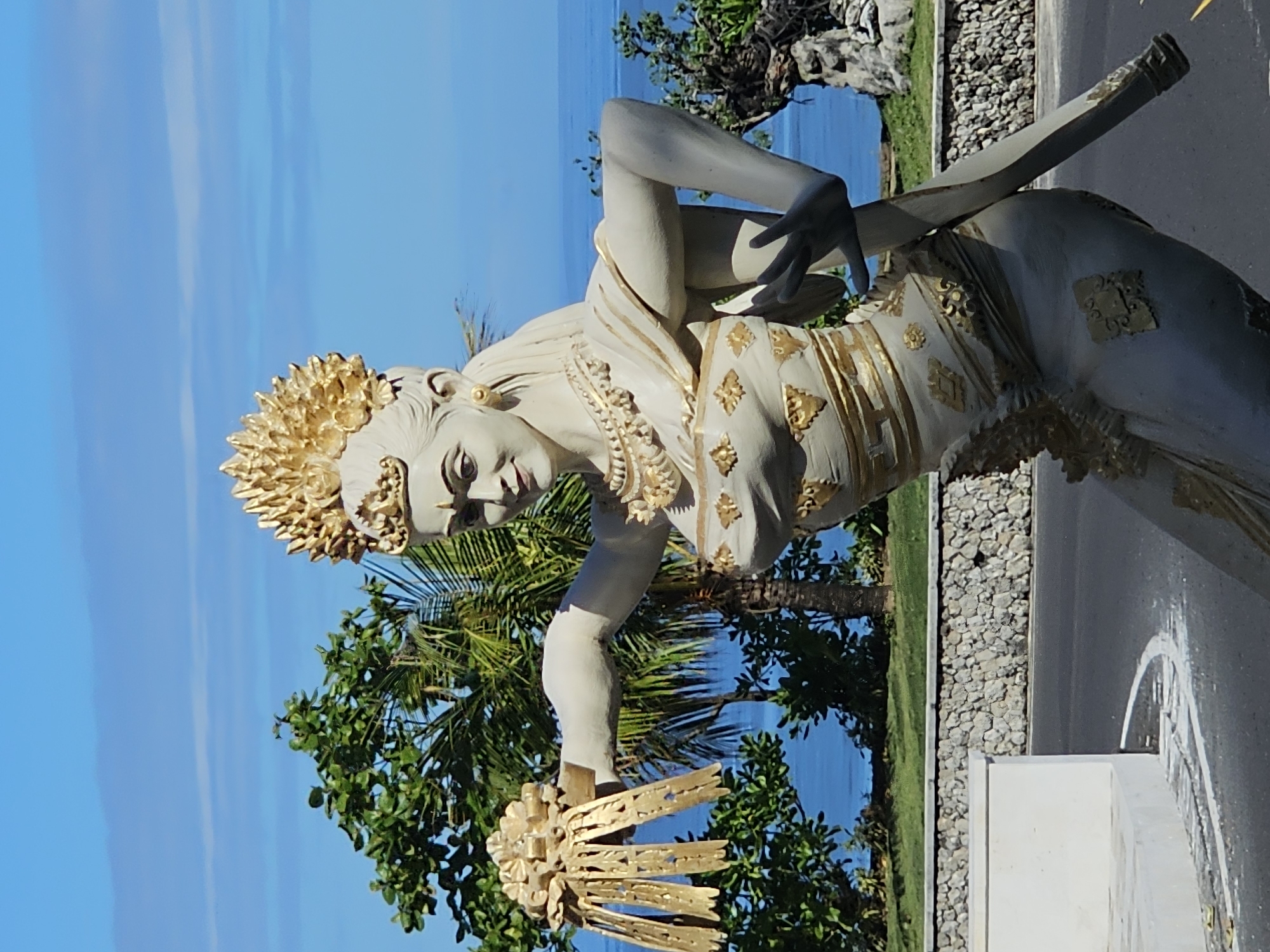 The image depicts a stone statue of a Balinese dancer in Kuta Selatan, Indonesia. The statue is adorned with gold detailing on its headdress, dress, and hand ornament. The dancer strikes a pose with one arm raised and the other lowered, holding the golden ornament. Behind the statue, the ocean and blue sky are visible. Trees and a road with a stone-lined border are also present.
