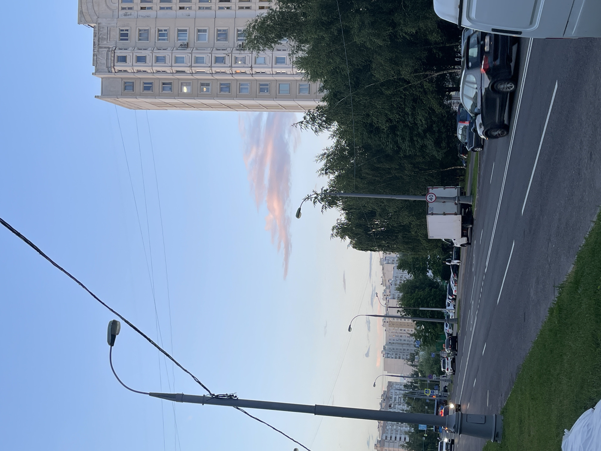 The image captures a street scene in Moscow, Russia, under a twilight sky transitioning from blue to pink. A tall, modern apartment building with numerous windows is visible on the right, partially obscured by a dense line of dark green trees. A street light with a curved arm stands prominently on the left side of the frame. The road in the foreground features multiple lanes of traffic. Several vehicles, including cars and a white truck, are present. In the background, a row of apartment buildings stretches across the horizon. The overall ambiance suggests an urban setting at dusk.