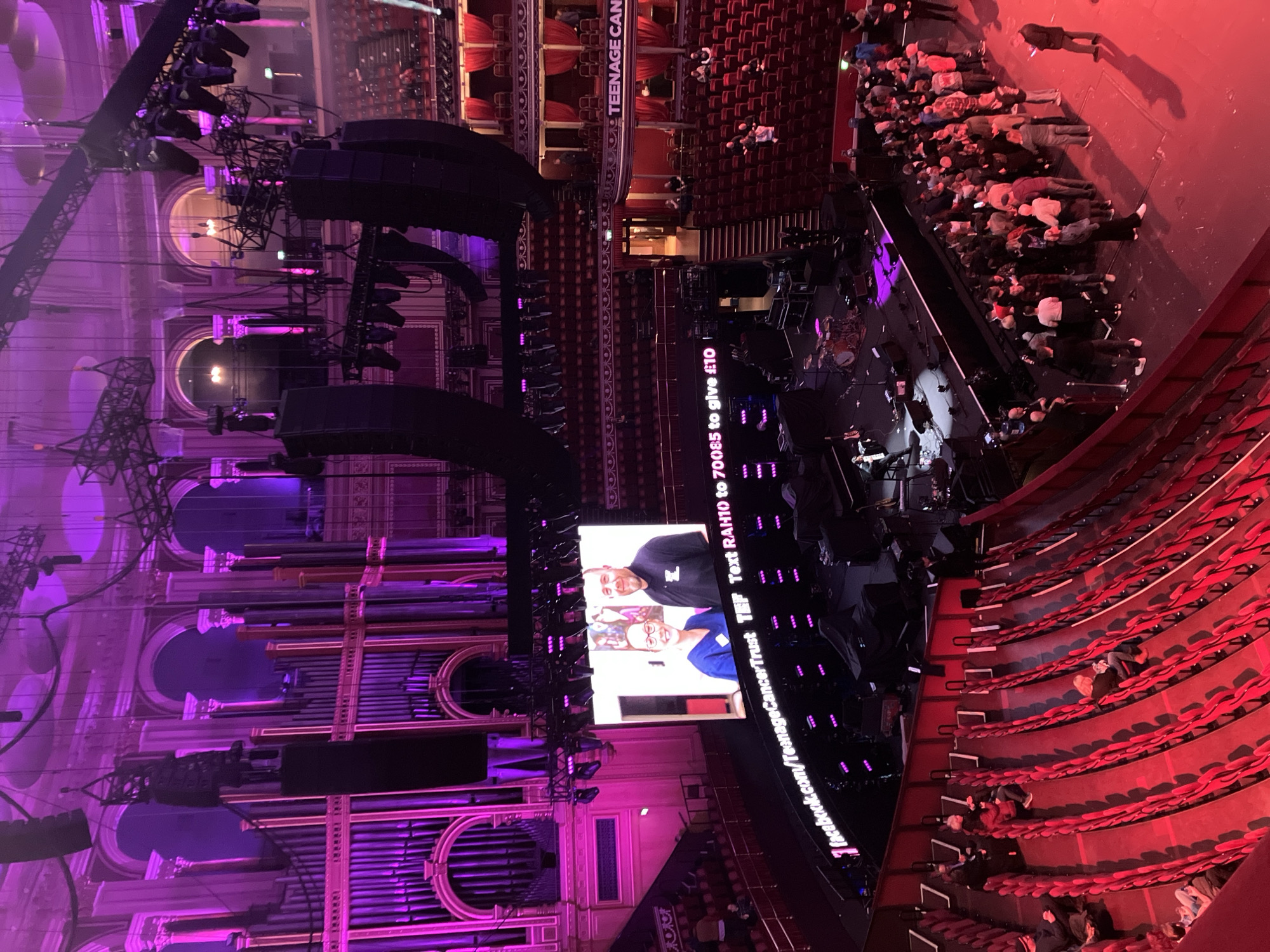 The image depicts the interior of the Royal Albert Hall, Kensington Gore, London, United Kingdom, seen from an elevated position within the upper tiers. The large, circular concert hall is illuminated predominantly with deep purple and magenta lighting, casting a dramatic glow on the ornate classical architecture, including arches, columns, and a prominent pipe organ structure on the left side. Red upholstered seating occupies multiple tiers, with most seats vacant, though scattered individuals and small groups are present in various sections.
The central stage area features a black grand piano with a single individual seated, performing or rehearsing. A drum kit is partially visible to the left of the piano. Numerous large black speaker arrays are suspended from the ceiling by metal trussing, accompanied by extensive lighting rigs that contribute to the purple illumination.
A large central LED screen prominently displays an image of two smiling individuals (a woman in medical scrubs and a man in a black t-shirt) above the text "TeenageCancerTrust" and "Text RAH10 to 70085 to give £10". Below this, a horizontal LED ticker displays "facebook.com/TeenageCancerTrust".
On the floor level to the right of the stage, a crowd of approximately 70-100 people is gathered in a loose queue or congregation. Individuals within this group are dressed in casual attire and appear to be waiting. A few staff or security personnel are visible near the stage and within the seating areas. The overall scene suggests a charity event or concert in its preliminary stages.