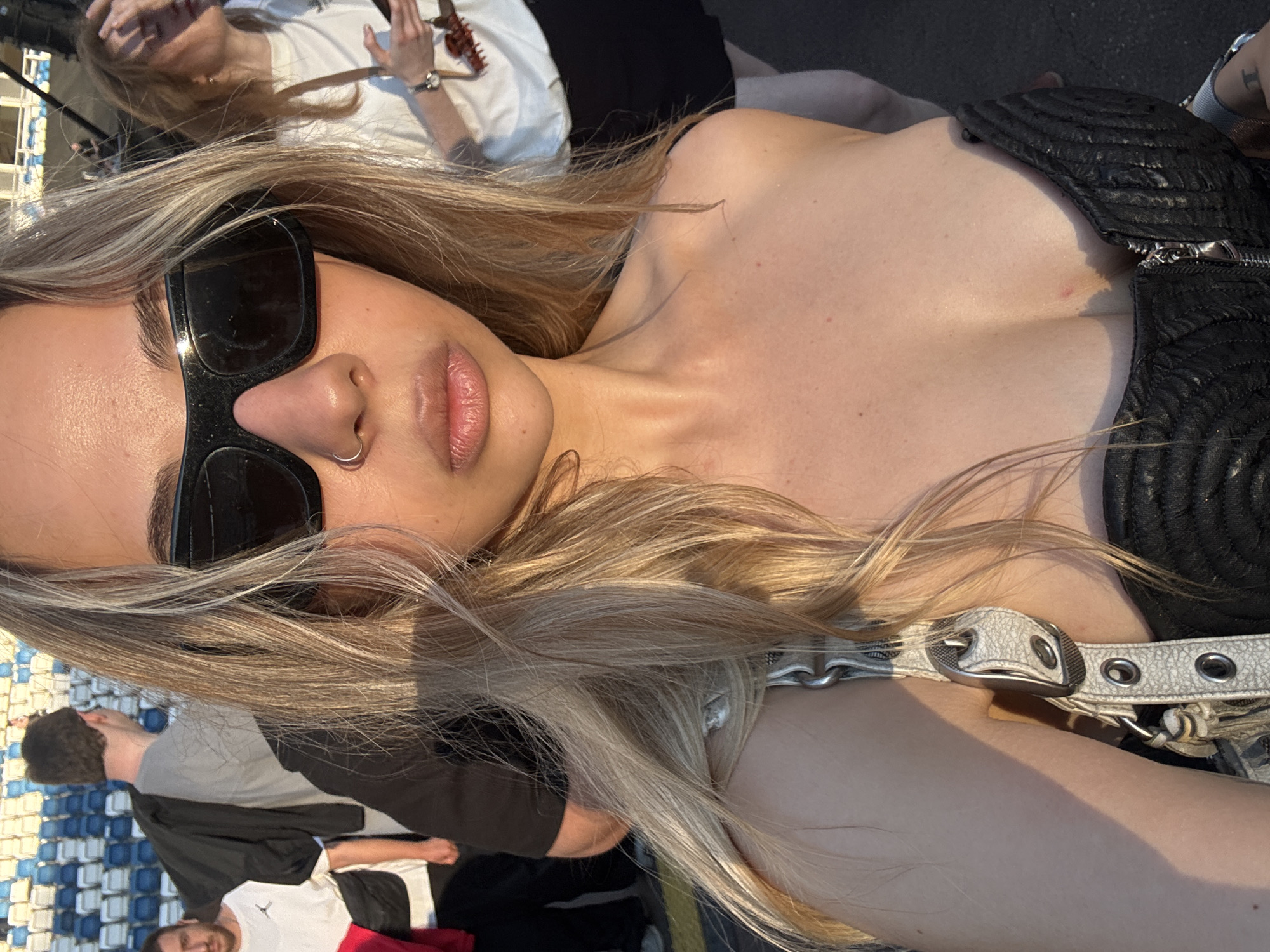 A woman with blonde hair and sunglasses takes a selfie in an outdoor urban setting. She is wearing a black strapless top and a white purse with a buckle strap. In the background, there are bleachers, people standing and walking, and a stage. The image is taken during the day and the lighting suggests it might be late afternoon. The location is not specified.
