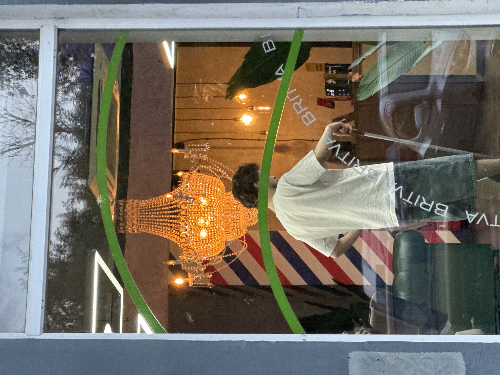 The image captures the interior of a barbershop or salon, viewed from outside through a window at what appears to be evening or night. A man with dark hair, wearing a white t-shirt and dark shorts or trousers, stands with his back largely to the viewer. His right arm is raised, holding a long, slender, metallic object, possibly scissors or a comb.
Inside, a large, ornate crystal chandelier with numerous lightbulbs hangs brightly from the ceiling, casting a warm, golden glow. Behind the man, a distinctive barber pole pattern of diagonal red, white, and blue stripes is visible on a wall or screen. Several bare light bulbs on wires are strung across the ceiling, contributing to the ambient lighting. Large green leaves, possibly artificial plants, are seen in the upper portion of the interior, near the ceiling. A dark object, which could be a barber chair, is partially visible towards the bottom right.
The window through which the scene is viewed reflects the outside environment, including bare tree branches, suggesting a winter or late autumn setting, along with glimpses of a light-colored building and the sky. A bright green, curved strip is adhered vertically to the window glass, acting as a decorative element.
The word "BRITVA" (Russian for "razor") is prominently displayed in white capital letters on the interior elements or as applied vinyl on the window, with partial letters like "B" and "A" also visible. The overall atmosphere is inviting, with the bright interior lights contrasting with the darker exterior.