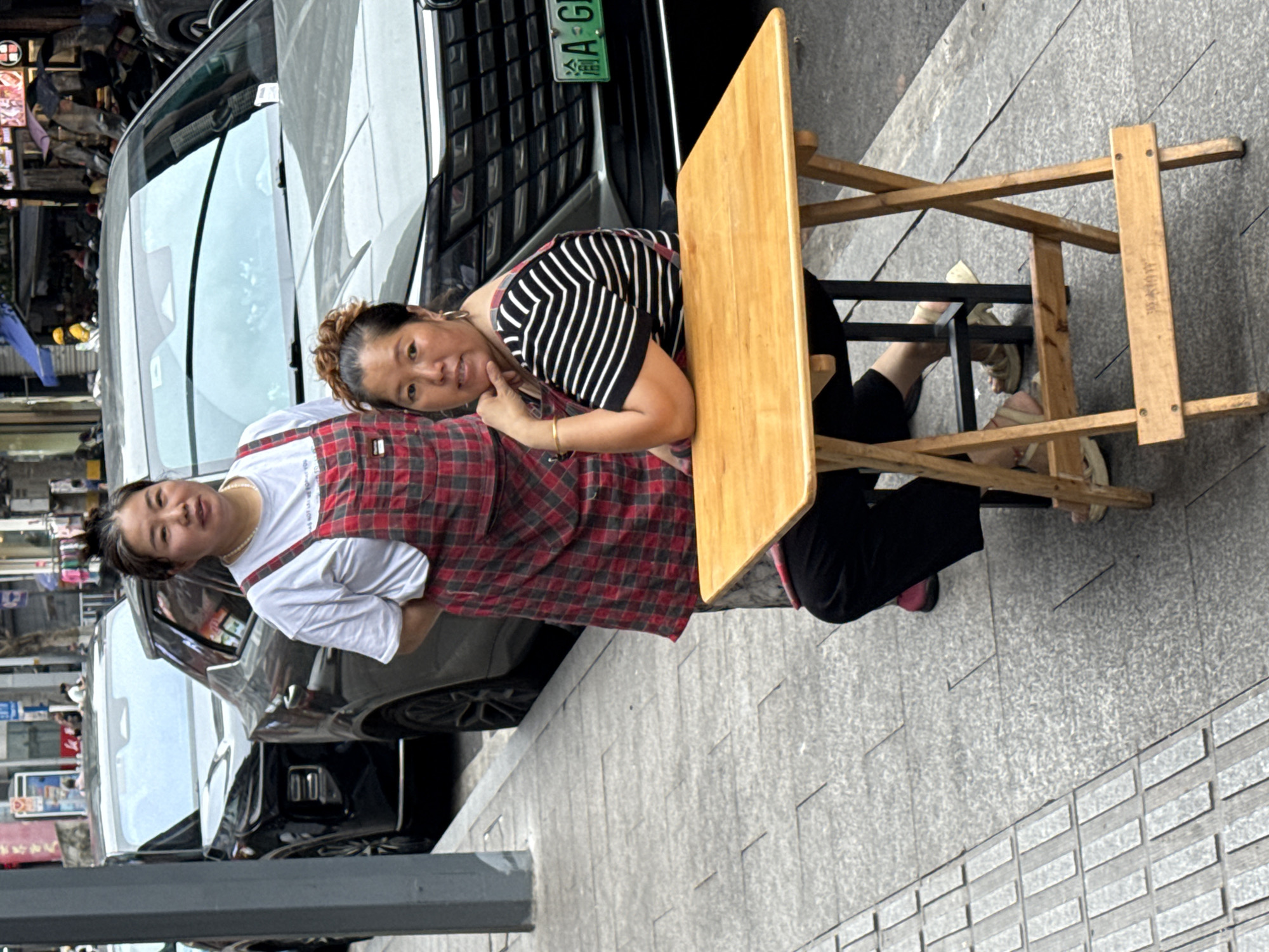 Two Asian women are positioned at a small, light-brown wooden folding table set up alongside a parked black car on a city street in Jiangjin, China. One woman is leaning on the table with her chin on her hand, wearing a black and white striped shirt and black pants. The other woman is standing behind her wearing a white shirt and a red plaid apron. The sidewalk is paved with rectangular gray stones. Shops and other cars are visible in the background on the other side of the street. A green license plate is visible on the parked car.