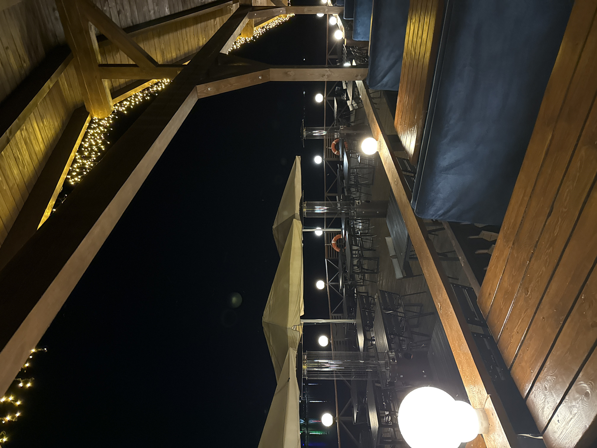 The image depicts an outdoor dining area at night, situated in Zolotoi Plyazh, Russia. The scene is illuminated by string lights draped along the wooden beams of a covered structure, and by spherical lamps placed around the perimeter of the deck.
The foreground features wooden tables and dark blue upholstered benches, suggesting a seating area for patrons. In the mid-ground, there are numerous other tables and chairs set up, some covered by large, light-colored umbrellas. Several outdoor heaters are visible between the tables, indicating that this space is likely used in cooler weather. A bright, white spherical lamp is prominent on the left side of the image, casting a strong glow on the adjacent wooden railing and table.
Beyond the dining area, a railing separates the deck from a body of water, which appears to be a calm lake or sea. Lights are visible in the distance on the far side of the water, reflecting on its surface. The overall atmosphere is serene and inviting, set against a dark night sky. There are no people visible in the image.