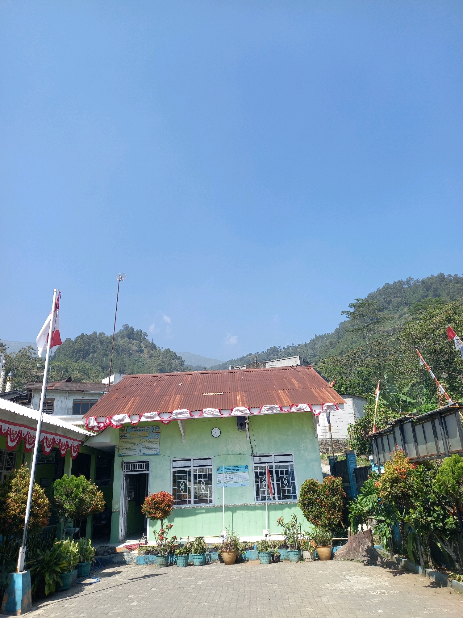 The image captures a building with a light green facade, likely a government office or community center, situated in the Kebaturan area of Indonesia. The building features a rusty corrugated metal roof adorned with red and white festive bunting. Several windows with white frames are visible, some decorated with small Indonesian flags. In front of the building, a paved courtyard is lined with potted plants and trees. A flagpole with a red and white Indonesian flag stands prominently to the left of the building. In the background, a lush green hillside covered in trees rises under a clear blue sky. The overall impression is one of a well-maintained public space in a rural or semi-rural setting.