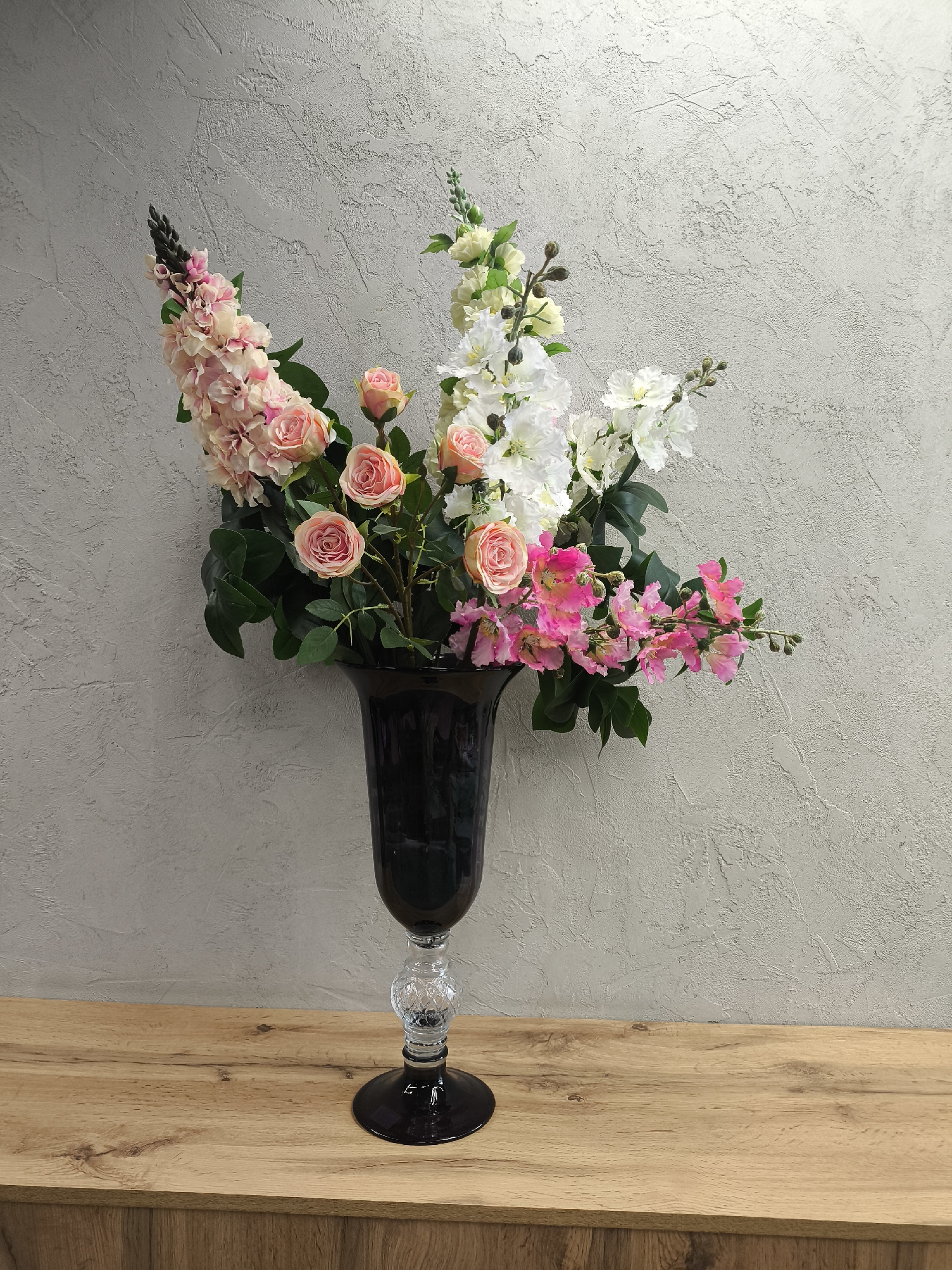 A black glass vase filled with assorted pink and white flowers is centered on a wooden surface against a textured gray wall. The vase has a clear, patterned stem. The flowers include roses, stock, and other varieties. The wooden surface appears to be part of a cabinet or table. The image provides no information indicating it was taken in Izhevsk, Russia.