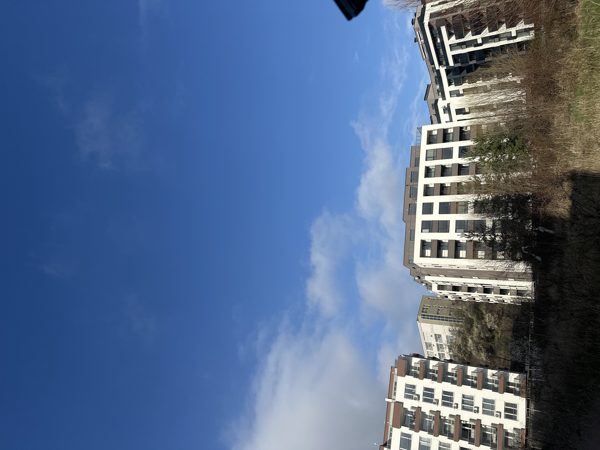 The image depicts a low-angle view of several modern residential buildings in Sofia, Bulgaria, juxtaposed against a clear, blue sky with scattered, light clouds. The buildings feature predominantly white facades with brown or gray accents and visible balconies or window frames. The surrounding area appears to be partly vegetated with trees and shrubs, some exhibiting bare branches, suggesting it is either late autumn or early spring. There are no people visible in the shot. The buildings appear to be of modern architectural style and construction.