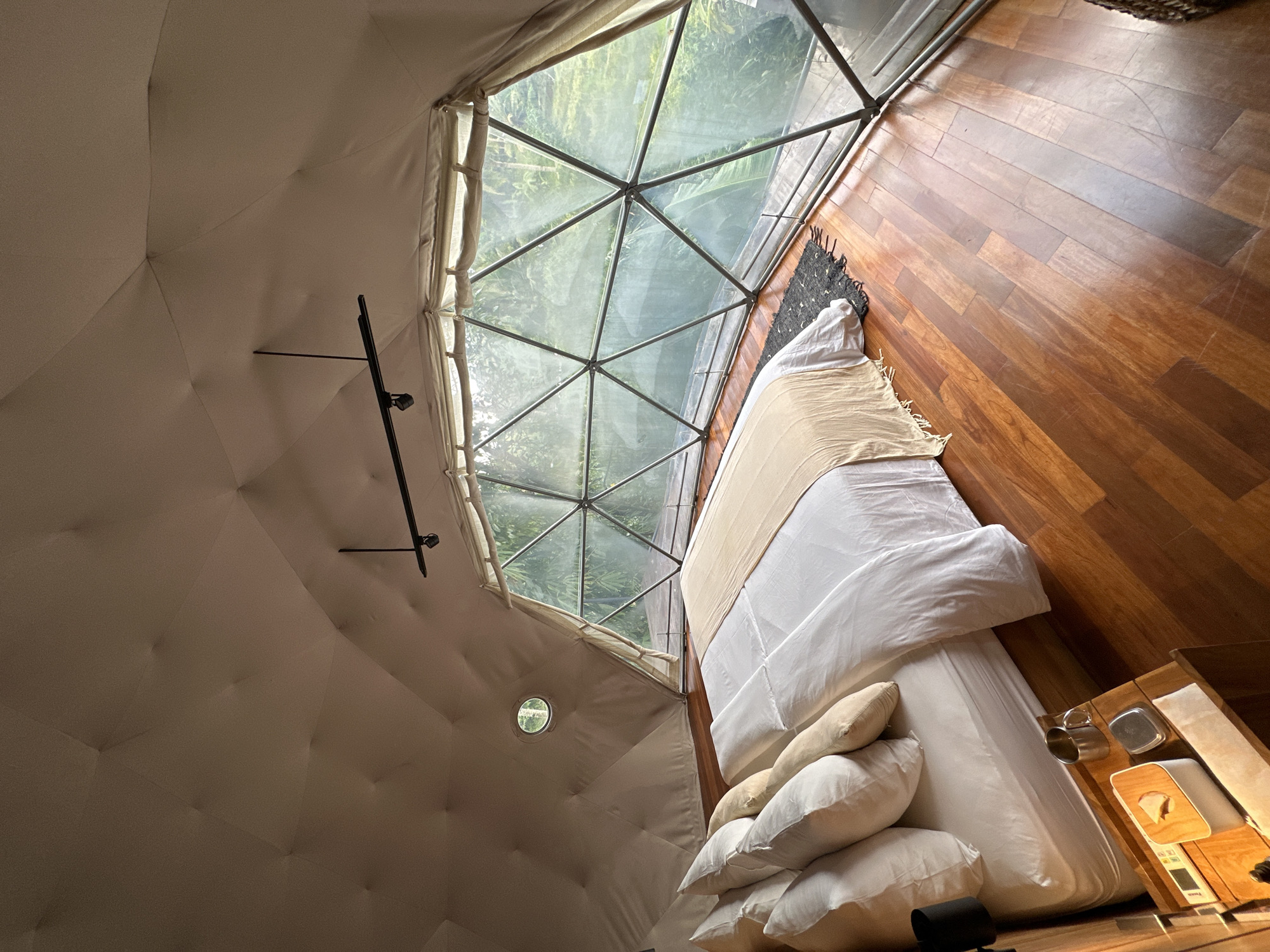 The image depicts the interior of a geodesic dome structure, possibly a glamping site, located in Licin, Indonesia. The dome features a large trapezoidal window composed of triangular glass panes offering a view of lush greenery, likely the surrounding forest. The interior is dominated by a large bed with white linens, several pillows, and a tan throw blanket. A small wooden bedside table is positioned next to the bed, holding various items including containers and a tissue box. The floor is made of polished wood planks. The walls of the dome are covered in a light-colored fabric, and a black metal rod, likely for hanging items, is mounted on one section of the wall. A small, circular window is also visible on the dome wall. The overall atmosphere is one of comfort and immersion in nature.