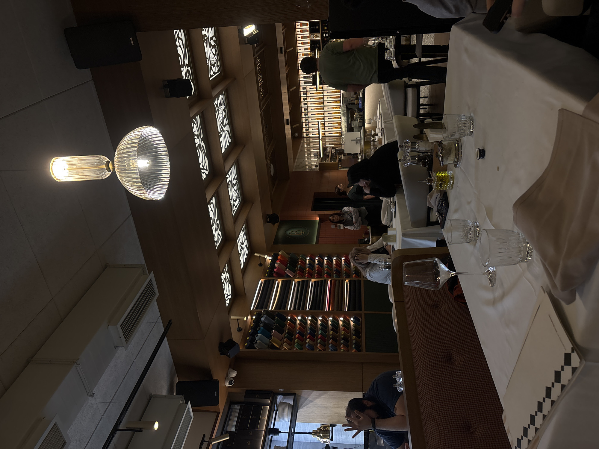 This media file captures the interior of what appears to be an upscale restaurant, bar, or a combined retail/lounge space, likely in the evening given the dim, artificial lighting. The ambiance is warm and modern, characterized by wooden walls, illuminated decorative ceiling panels with white patterns, and strategic lighting.
Several individuals are present, suggesting social activity. In the mid-ground, a person in a green t-shirt stands near a counter, while another person is seated at a table covered with a white tablecloth, set with crystal glasses and condiment containers. In the lower foreground, a bearded man in a dark shirt is leaning on a surface. Other patrons are visible further in the background, some seated near a distinctive shelving unit.
A prominent feature is a large wooden shelving unit along a back wall. It displays numerous colorful items, meticulously arranged in rows, which could be spools of thread, small bottles, or cigars. Above these, neatly stacked rolls of fabric or folded garments are visible. A green sign with a stylized, indistinct logo is hanging on the wall near these shelves.
The lighting includes a unique ribbed glass pendant lamp prominently featured in the foreground, along with various recessed and strip lights. Overhead, black speakers and white ventilation ducts are mounted on the ceiling. A security camera is also visible. On one of the tables, a white card with a black and white checkered pattern along its edge is placed next to the glassware. The overall setting suggests a contemporary and refined commercial establishment.