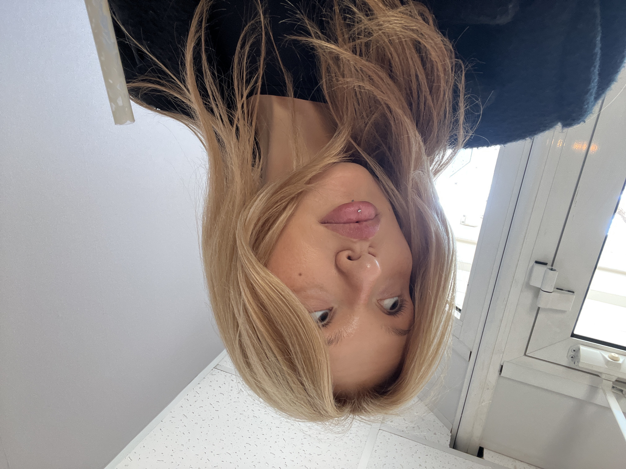 A woman is depicted in an upside-down view, with a piercing visible on her tongue. She has blond hair and fair skin. She is wearing a dark-colored garment. The environment appears to be indoors, near a window, and a ceiling with a grid pattern is visible. The image was taken in the city of Chaikovsky, Russia.