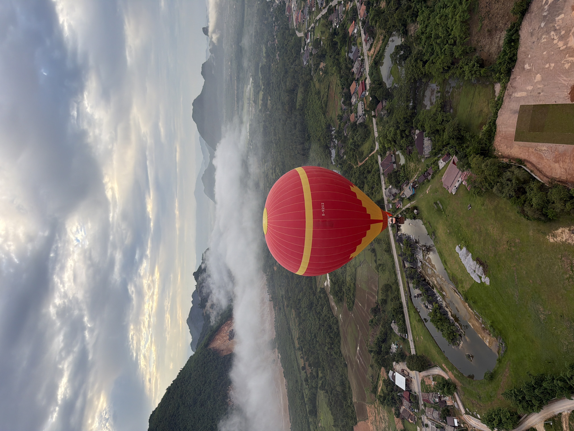 An aerial, wide-angle shot captures a vibrant red and yellow hot air balloon ascending over a misty, mountainous landscape at dawn. The balloon is in the foreground, dominating the right side of the frame. In the mid-ground, a village with scattered houses and green fields is visible, bisected by a narrow road. A winding river or canal cuts through the landscape, lined with trees and small buildings. The background features lush green mountains shrouded in a layer of mist, with darker, more distant peaks visible on the horizon. The sky is overcast with diffused sunlight breaking through the clouds. The scene appears to be Hong Ngern Village, Laos.