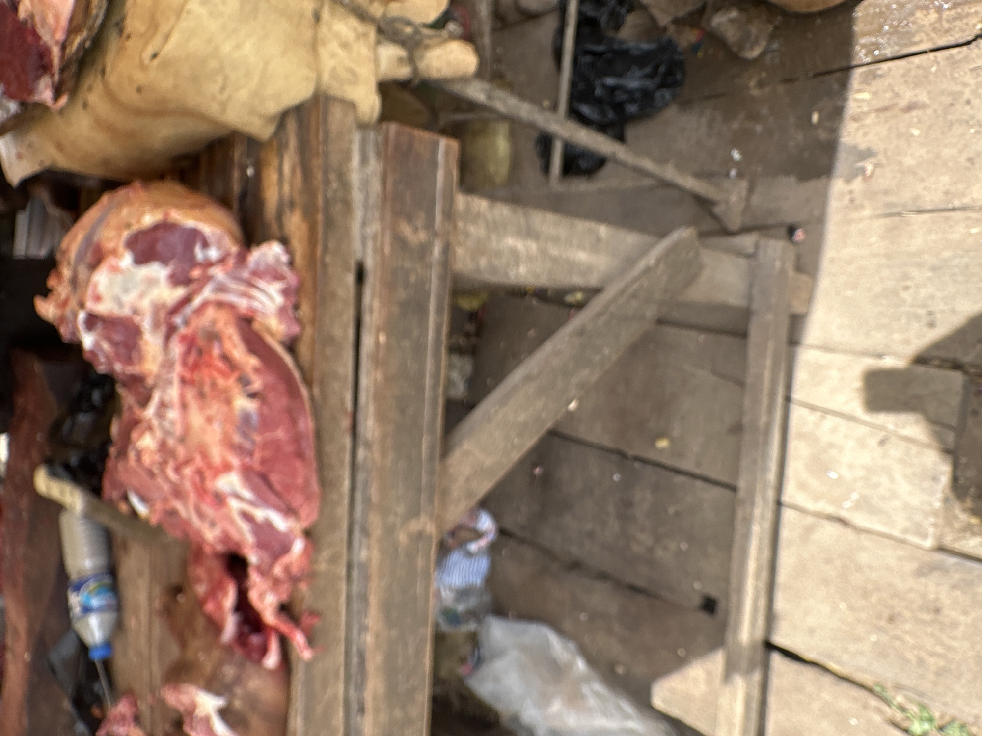 The image depicts a butcher's stall in Narayi, Nigeria. On a wooden table, large cuts of raw meat are visible, some with bone and fat. A knife is partially embedded in a piece of meat. To the left, a plastic bottle of water rests on the table. The area beneath the table is cluttered with discarded materials, including a plastic bag and what appears to be a striped fabric. The ground is made of wooden planks. The scene is lit by natural light, casting shadows.