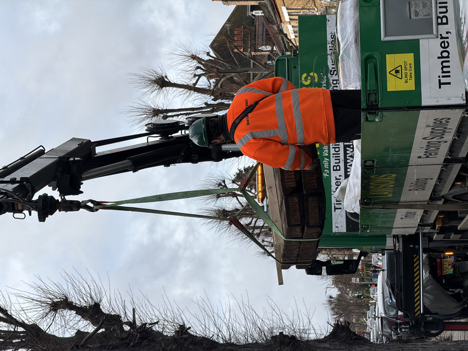 The image depicts an outdoor urban scene under an overcast sky, showing a commercial delivery operation. A male worker, viewed from behind, wears an orange high-visibility jacket with reflective stripes, black trousers, and a dark green hard hat. He stands on the flatbed of a green and white 'Lawson's Timber, Building, Fencing Supplies' truck. A hydraulic crane arm, mounted behind the truck's cab, is extended and actively lifting a bundle of dark brown, stacked timber materials, possibly railway sleepers, secured by a green synthetic sling. An orange flashing beacon is active on the crane arm. The truck's registration plate, 'LG72 CWE', is visible, identifying it as a UK vehicle. Bare, pollarded trees line the street, and typical London residential brick buildings with pitched roofs are visible in the background. A line of parked vehicles is present along the street, indicating an active thoroughfare. The scene is located on an urban street in London, United Kingdom.