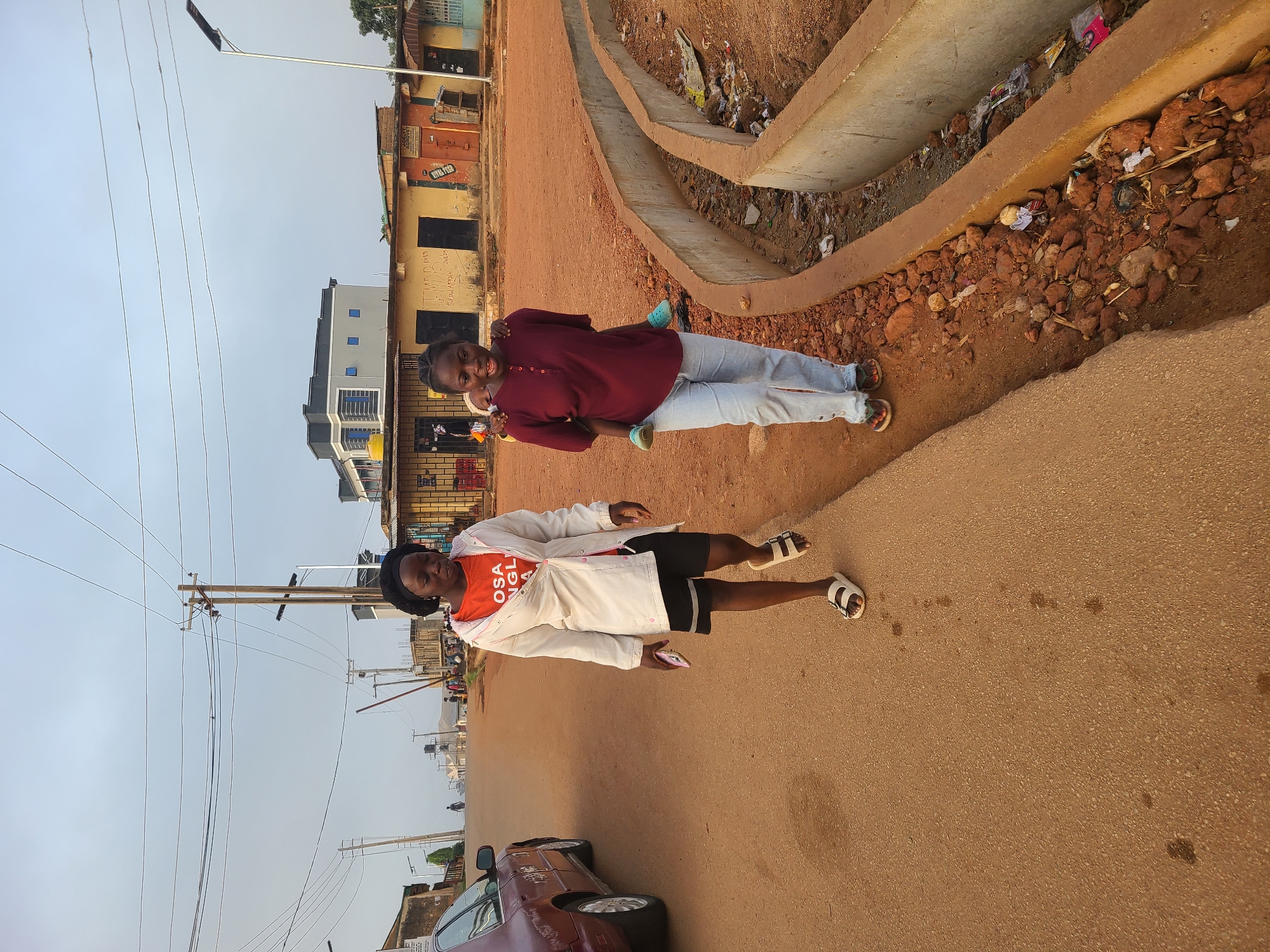 Two women are standing on a street in Jos, Nigeria. One woman is giving the other a piggyback ride. The woman giving the ride is wearing a long-sleeved, red top, light-colored pants, and sandals. The woman receiving the ride is wearing an orange t-shirt, a white jacket, black shorts, and white sandals. She is holding a phone. A car is parked on the side of the road. A concrete gutter runs along the side of the street, filled with debris. Buildings are visible in the background. Power lines run above the street. The sky is overcast.