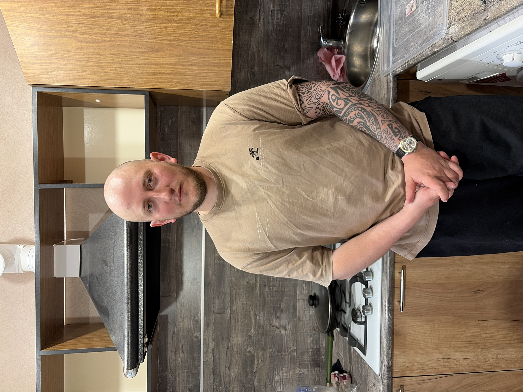 A man stands in a kitchen. He has a shaved head and a beard. He is wearing a tan t-shirt and black pants. His left arm is covered in tattoos. He is wearing a watch on his left wrist. He has his hands clasped in front of him. Behind him is a kitchen with a sink, a stove, and cabinets. The cabinets are a combination of light brown wood and dark gray. There is a range hood above the stove. It's unclear if this location is in the city of Izhevsk, Russia.