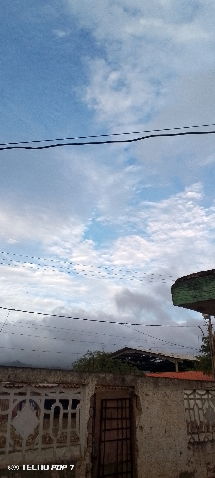 The image shows a view of the sky and a street scene in Altagracia, Venezuela. The sky is partly cloudy with blue patches visible. Several power lines are strung across the frame. In the foreground, there is a weathered wall with a decorative white fence on the left and a metal gate. Behind the wall, a building with a damaged roof and an orange-tiled roof is visible. A green-covered structure is partially visible on the right, possibly a bus stop or shelter. The overall impression is of a somewhat dilapidated urban environment under an overcast sky.