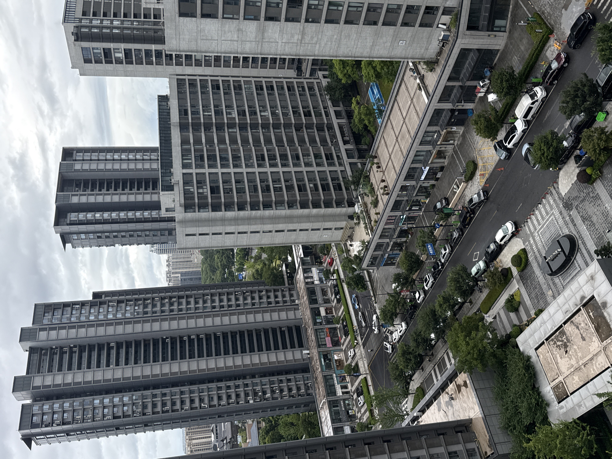 The aerial image depicts a modern urban landscape in Yiwu, China, characterized by several high-rise residential buildings and commercial structures. The buildings are predominantly gray and feature repetitive window patterns, indicative of contemporary architectural design. In the foreground, a street is lined with parked cars, including sedans, SUVs, and vans. The street also shows pedestrian walkways and green spaces with trees and bushes. Several commercial storefronts are visible along a low-rise building adjacent to the street. Further in the background, more residential towers dominate the skyline, some partially obscured by trees. The sky is overcast with clouds. There are no discernible interactions between individuals visible in the scene, and the overall impression is one of a developed, bustling urban environment.