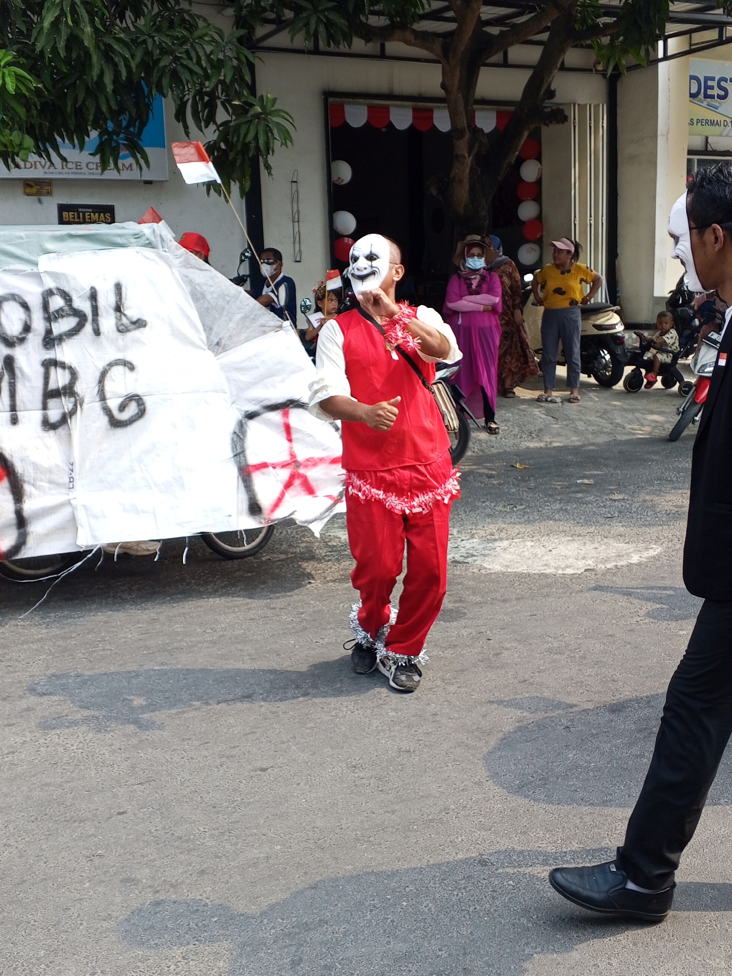The image depicts an outdoor street scene, likely in Ciruas, Indonesia, possibly during a parade or event. In the foreground, a person in a red costume with a white clown-like mask is standing, with their body angled towards the right. They are wearing red pants and a red vest over a white shirt, with white fluffy trim around their neck and ankles. The person appears to be gesturing with their right hand. To the right of this person, another individual in a black suit and black shoes is partially visible, facing away from the camera.
In the background, a white, tent-like structure or vehicle adorned with black lettering and red markings is situated on the left side of the frame. A small Indonesian flag is also visible near this structure. Several other people are present, including a woman in a pink dress and a headscarf, and another woman in a yellow shirt and pink cap. A child is seated on a small, motorized vehicle behind them. Scooters and a building with signage are also visible in the background. The ground is paved, and shadows are cast from the people and objects, indicating it is daytime. The environment suggests an urban or semi-urban setting with trees and buildings.