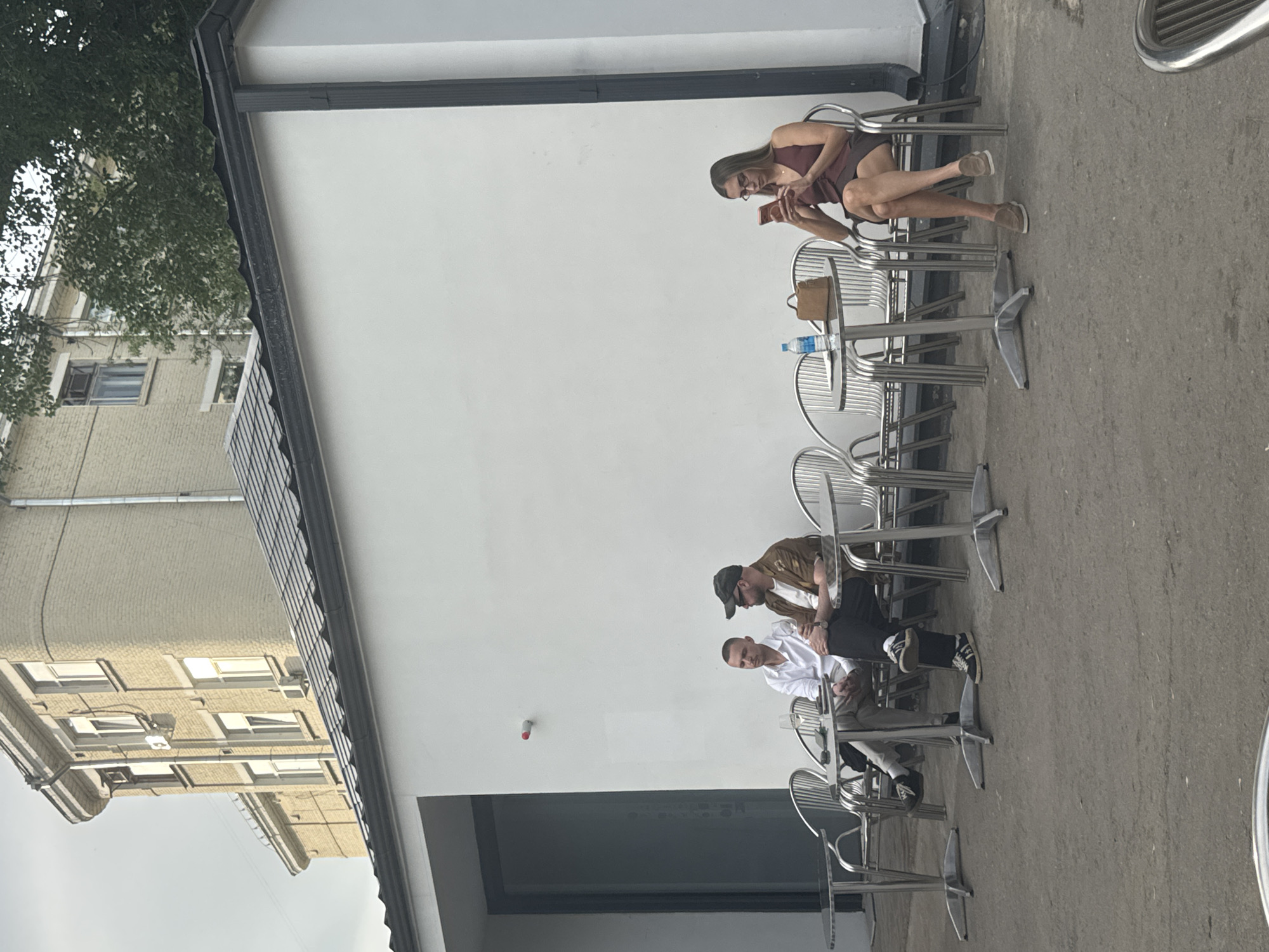 The image shows an outdoor scene. Three individuals are seated at separate tables, engaging in activities such as using a phone, conversing, and observing. The tables are arranged linearly against a white building exterior. The setting is likely an outdoor seating area of a cafe or restaurant. The surrounding environment includes a building with windows and trees visible in the background. The location cannot be determined as Moscow, Russia.