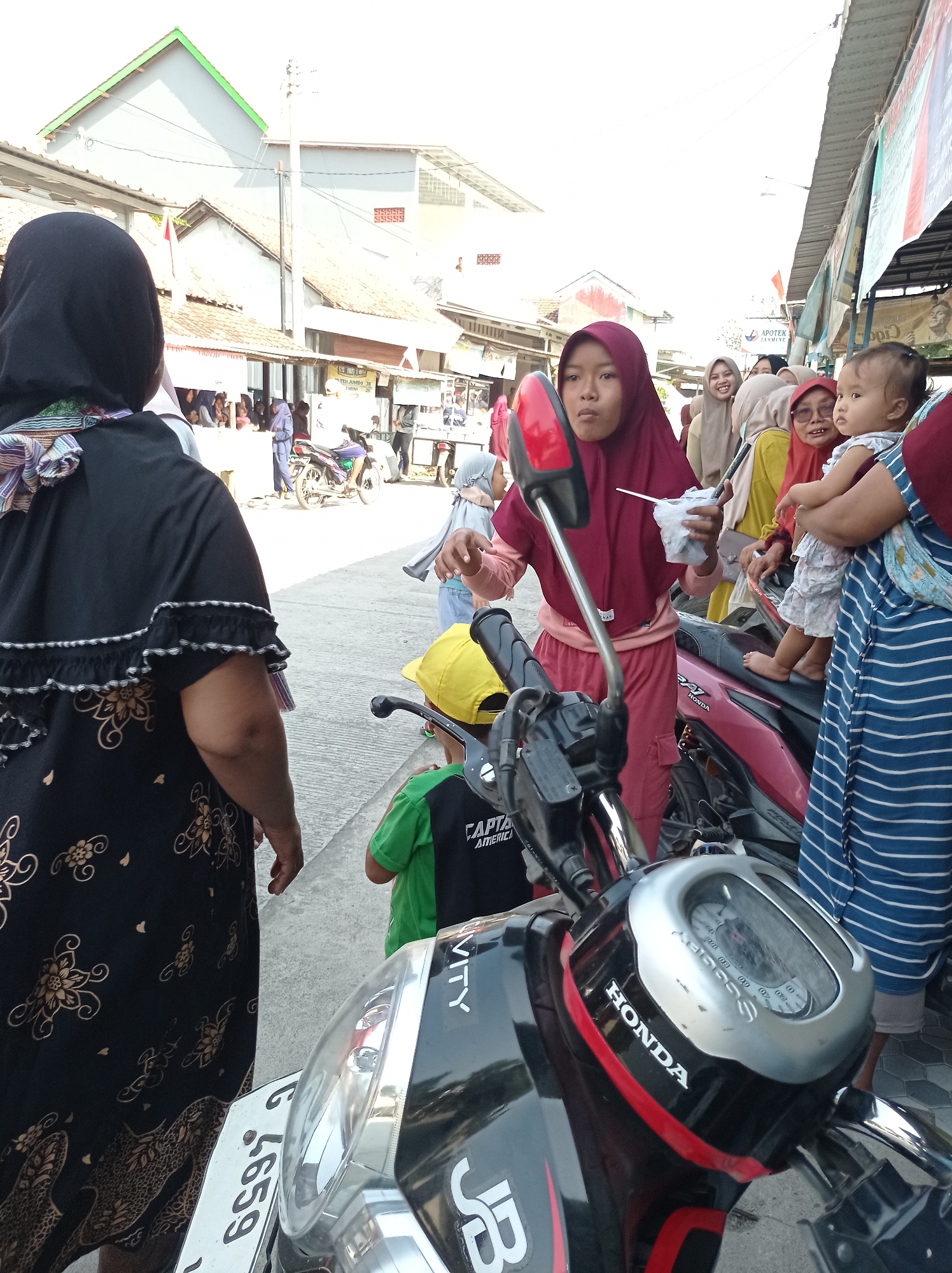 The image depicts a street scene in Maribaya, Indonesia. In the foreground, a portion of a black motorcycle is visible, with its silver and gray dashboard displaying some dials and the "HONDA" logo. To the right of the motorcycle, a person wearing a blue and white striped shirt stands next to a baby. A young girl wearing a maroon hijab and a pink shirt is holding a white wrapper and looking towards the left. Behind her, several other people are present, including a woman with a light brown hijab and another woman wearing glasses and a yellow shirt. In the background, buildings with tiled roofs and a street lined with stalls suggest a market or commercial area. A red motorcycle is parked to the right of the young girl. The overall impression is that of a busy outdoor gathering.