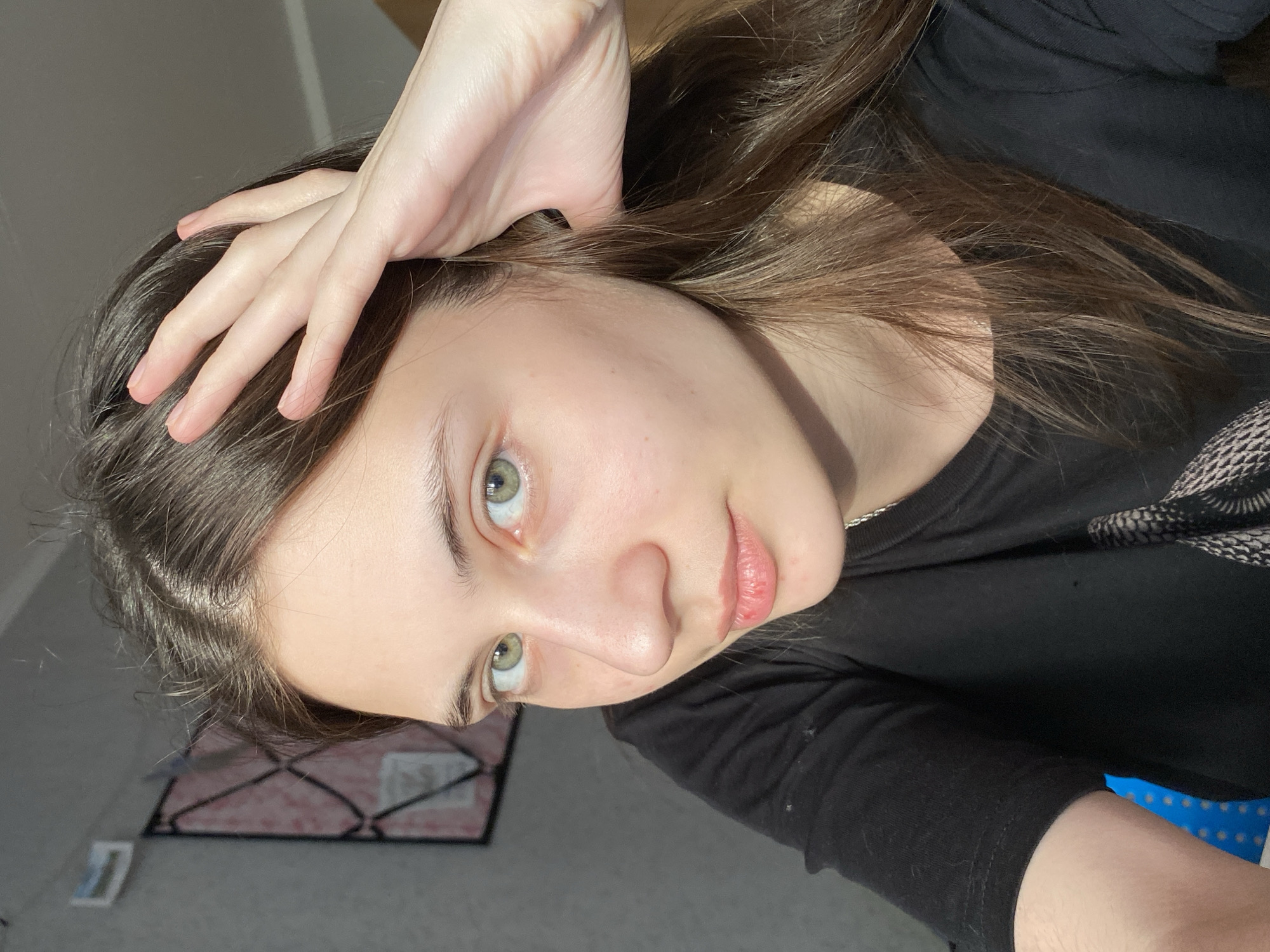 A young woman with fair skin and light brown hair is angled in the frame, her hand resting on her head. She has light green eyes and pink lips. She is wearing a black long-sleeved shirt. The background is out of focus but appears to be a room with a patterned object visible. There is no indication of Votkinsk, Russia.