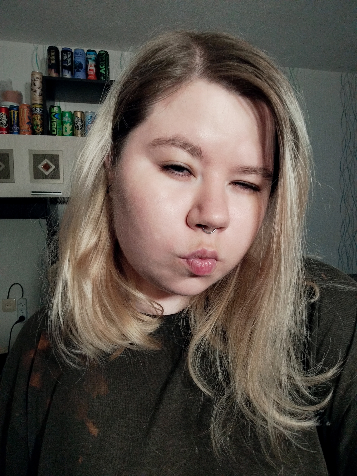 A medium close-up shot features a young adult with blonde hair and fair skin, winking with her right eye and pouting her lips. She has a septum piercing and is wearing a dark-colored t-shirt or sweatshirt with a subtle bleached pattern. The lighting suggests an indoor setting, possibly with artificial light.
In the background, to the left, is a wall-mounted shelf displaying a collection of various beverage cans. On the top shelf, from left to right, are cans with "1000ML" visible on several, and "VJOJIL" on a green can. On the bottom shelf, from left to right, cans are visible with text such as "coffe" (likely part of "coffee"), "MONSTER", and "ENERGY" on a green can, and "EVIGIL" on a blue can. Below the bottom shelf, two square framed artworks with diamond shapes are mounted on the wall. Further left, an electrical outlet with plugs connected is visible. To the right of the subject, the wall features a light-colored wallpaper with a subtle wavy pattern. The overall atmosphere conveyed by the subject's expression is playful.