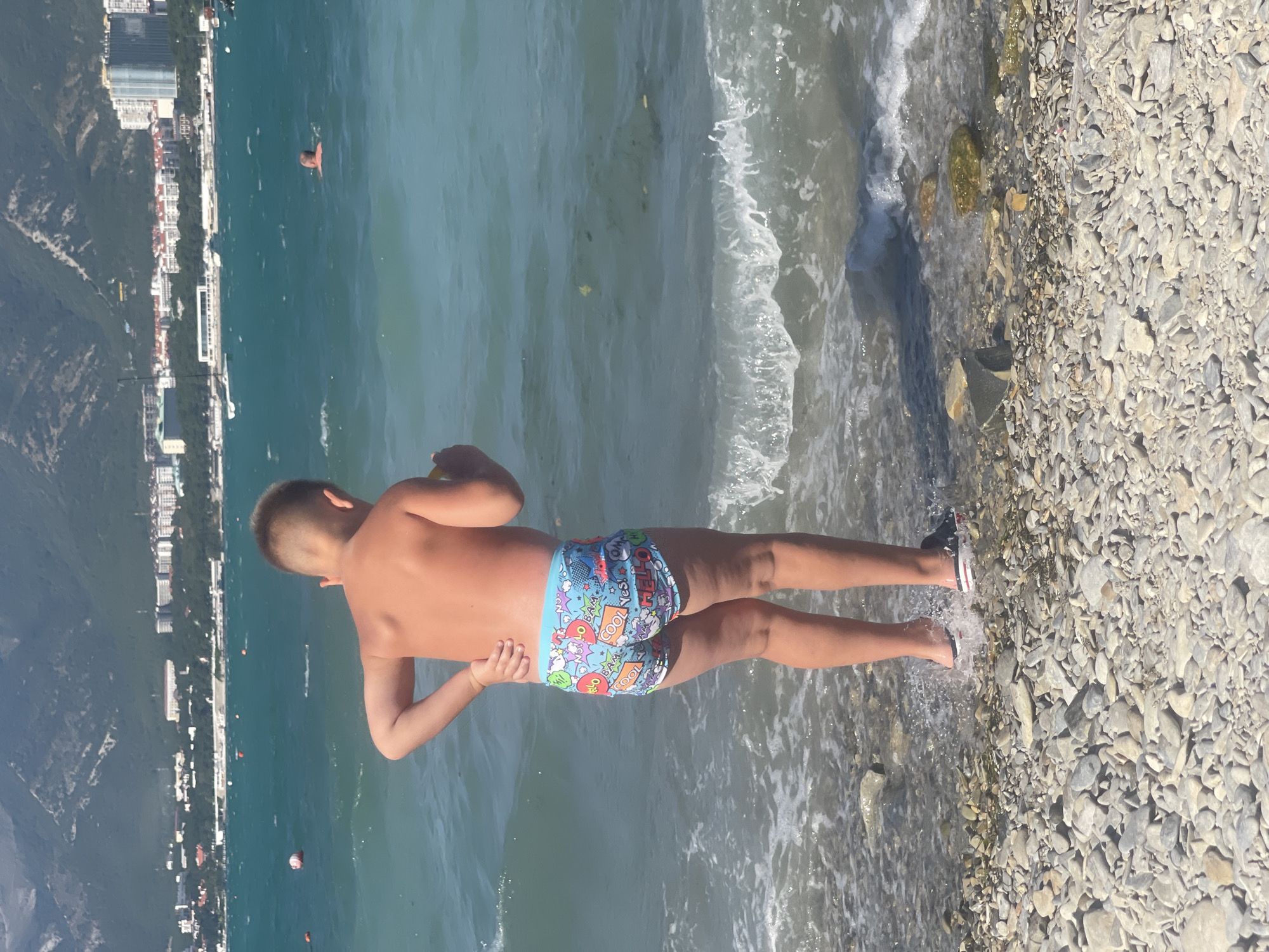 A young boy, facing away from the camera, stands at the edge of a pebble beach, his right hand on his lower back. He is wearing patterned swim trunks. Gentle waves lap at his feet, causing splashes. In the background, the turquoise water of the Black Sea stretches out, with a few buoys and a man swimming in the distance. A cityscape with numerous buildings and a backdrop of green, mountainous terrain is visible along the coastline. The sky is clear and blue. This scene is consistent with the coastline of Gelendzhik, Russia.