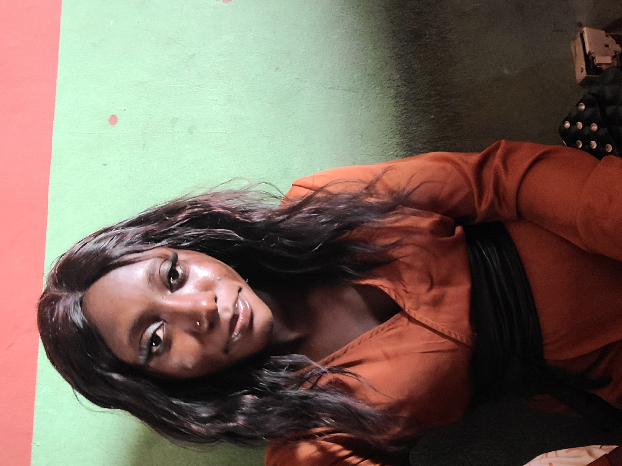 A young woman of African descent is pictured posing for a selfie in what appears to be an indoor setting with a textured green wall in the background. She has dark, wavy hair and is wearing a rust-colored V-neck top cinched at the waist with a black sash. Her makeup is noticeable, with emphasis on her eyes and lips. A small nose ring is visible. The lighting suggests it is likely daytime, possibly indoors with artificial light. The overall impression is a casual, personal photograph. The context of Gura Top, Nigeria, is not directly discernible from the visual elements in the image.