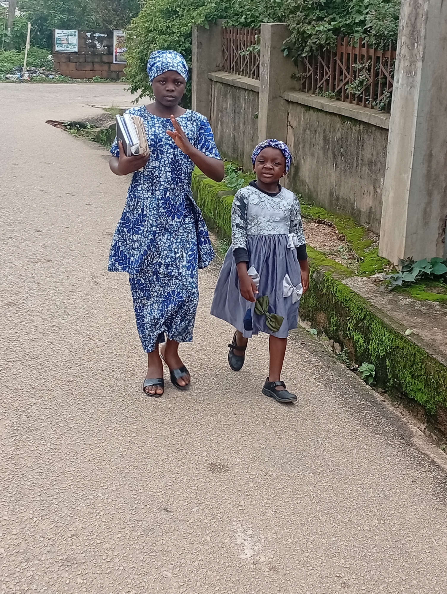 Two young girls walk down a paved road in an outdoor urban environment. The girl on the left is wearing a blue and white patterned dress and a matching headscarf. She is holding a stack of books in her left arm and raising her right hand slightly. The girl on the right is wearing a light gray dress with decorative bows and a blue patterned headscarf. She is looking forward with her mouth slightly open. A concrete wall with a metal fence runs along the right side of the road, covered in moss and vegetation. The road surface is asphalt. In the background, there are trees and a brick structure with posters.
