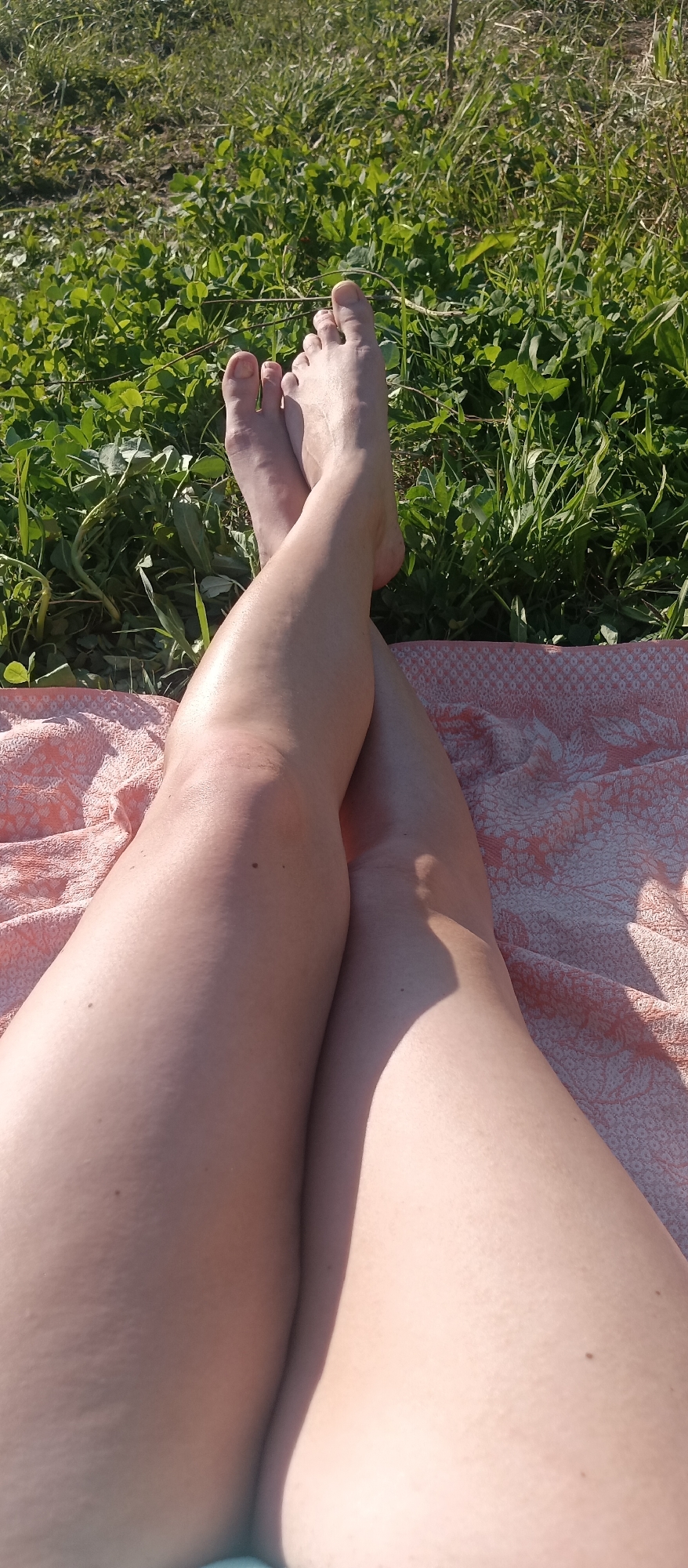 The image depicts a pair of crossed legs lying on a patterned blanket in a grassy outdoor setting. The legs are fair-skinned and have a few visible blemishes. The blanket is a light pink color with a pattern. The grass is green and lush. The location is in the city of Unknown, Unknown.