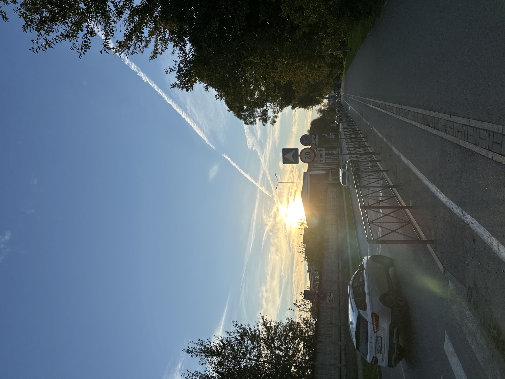 The image depicts a street scene at sunset in Perm, Russia. The sun is low on the horizon, casting a warm glow and long shadows across the asphalt road and adjacent sidewalk. A white car, identified by its license plate "H945YH 159", is driving on the road. The street is lined with trees and a metal fence separates the road from a pedestrian path. Traffic signs are visible, indicating a speed limit of 20 and a distance of 10 meters. The sky is a clear blue with wispy clouds and the contrails of an airplane.