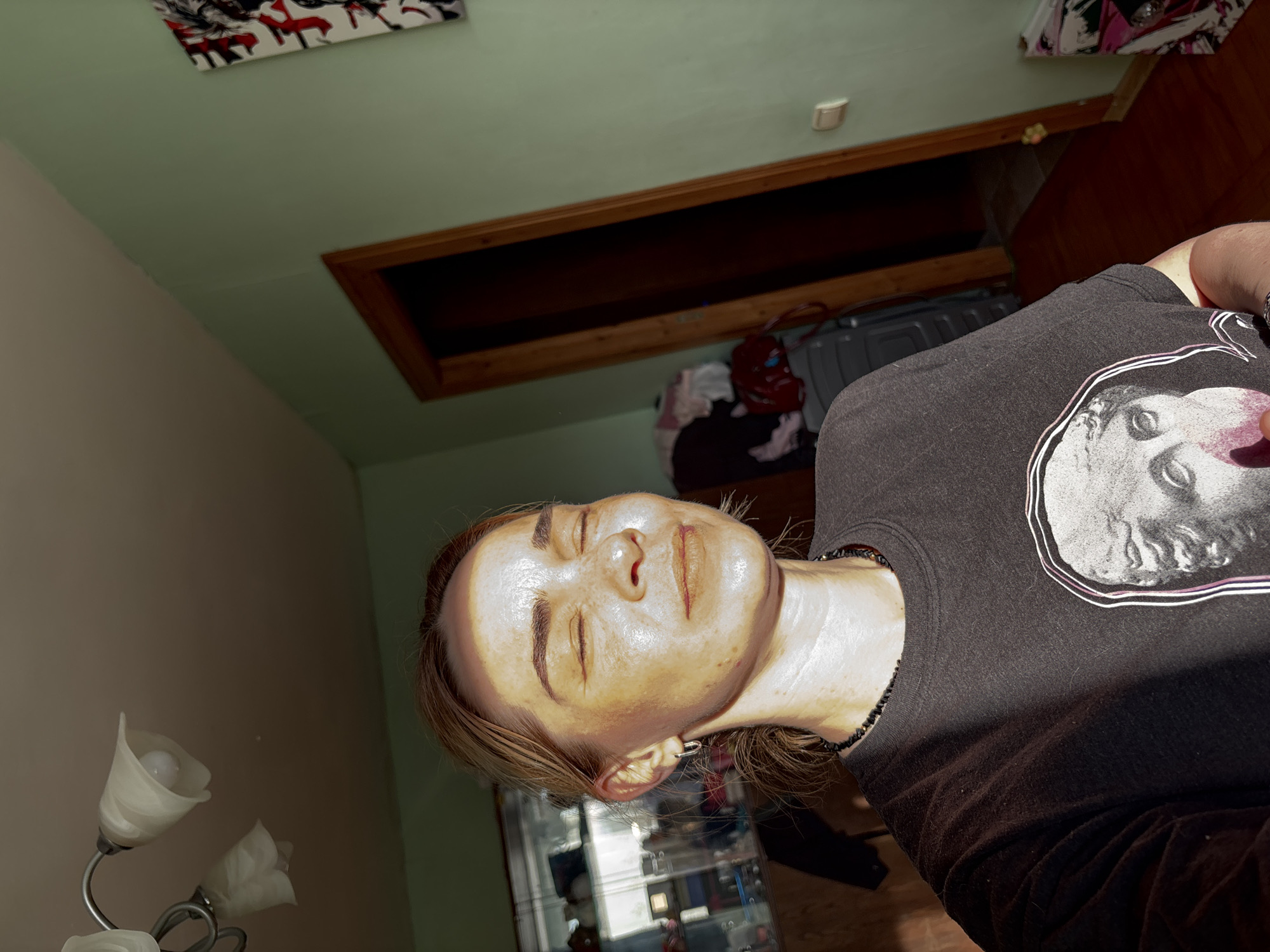 The image presents a tilted, low-angle view of a person lying down, seemingly undergoing a beauty treatment indoors. The subject is a female with light brown hair, eyes closed, and her face covered with a thick, shiny, yellowish facial mask. She wears a dark gray t-shirt adorned with a white graphic print of a classical bust, outlined with a thin purple line, and a black beaded necklace.
The setting appears to be a room with walls painted in two tones: a light green on the upper section and a lighter, possibly beige or off-white, on the lower section. A prominent feature is a rectangular attic hatch or storage opening in the wall/ceiling, framed by dark wood, with its interior being dark. Below this opening, several items are stored, including a red bag, folded clothing, and a gray plastic container. In the top left corner, a piece of wall art with red and white patterns is visible. A multi-armed lamp with white, tulip-shaped glass shades is seen in the lower left. Behind the person's head, part of a glass cabinet or shelving unit is discernible. The overall atmosphere suggests a relaxed moment in a home or residential setting in an unknown location in Russia. No discernible text is visible.