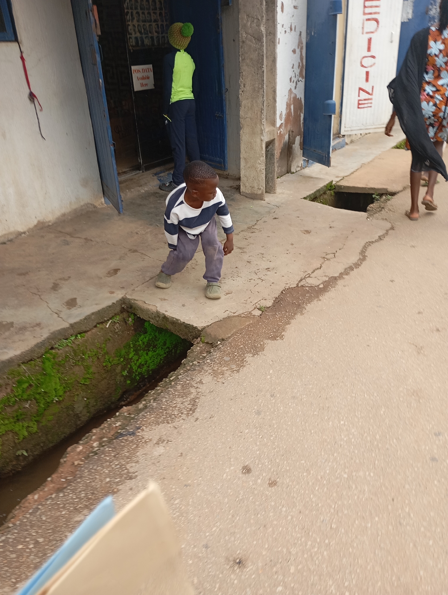 The image depicts a street scene in Jos, Nigeria. In the foreground, a young boy wearing a striped long-sleeved shirt and grey pants is walking on a cracked concrete sidewalk. To his left, a drainage ditch is visible, with green moss and some water. Behind the boy, a doorway is open, revealing a person wearing a bright green shirt and a yellow knitted hat. A sign on the wall inside the doorway reads "POS DATA Available Here." Further down the street, another individual, a woman wearing a patterned dress and a dark scarf, is walking away from the camera. The buildings on either side of the street appear to be commercial establishments, with blue doors and white walls. The street itself is paved with asphalt.