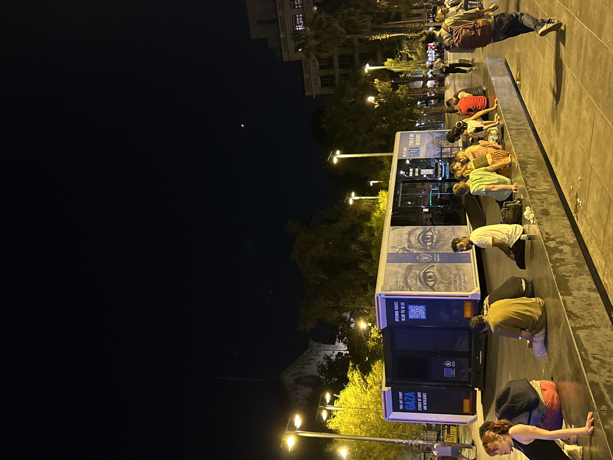 The image captures a nocturnal urban scene in Barcelona, Spain. In the foreground, a group of individuals are seated on a long, tiered concrete structure. To the left, a woman with red hair and a black top is leaning forward, her left arm extended. Beside her, another person, partially obscured, wears a black backpack. Further along the structure, several other people are seated, some in casual conversation.
Dominating the mid-ground is a large, mobile exhibition unit, resembling a trailer or bus, adorned with graphics. The signage on the unit clearly states "FREE ART EXHIBIT GAZA STORIES OF HOPE AND RESILIENCE" and displays "OPENING HOURS 10:00 TO 18:00" along with a QR code. The side of the unit features a large graphic of two eyes.
The environment suggests a public space, likely a plaza or walkway, illuminated by streetlights and the lights emanating from the exhibition unit and surrounding buildings. In the background, palm trees and other foliage are visible, along with architectural structures, including a building with multiple windows and a darker, more ornate structure with a steeple or tower. The sky is dark, indicating nighttime, with a faint celestial body visible. Several other individuals are walking in the background, and one person in a maroon backpack is walking away from the camera towards the right side of the frame. The ground surface is paved and shows scattered debris.