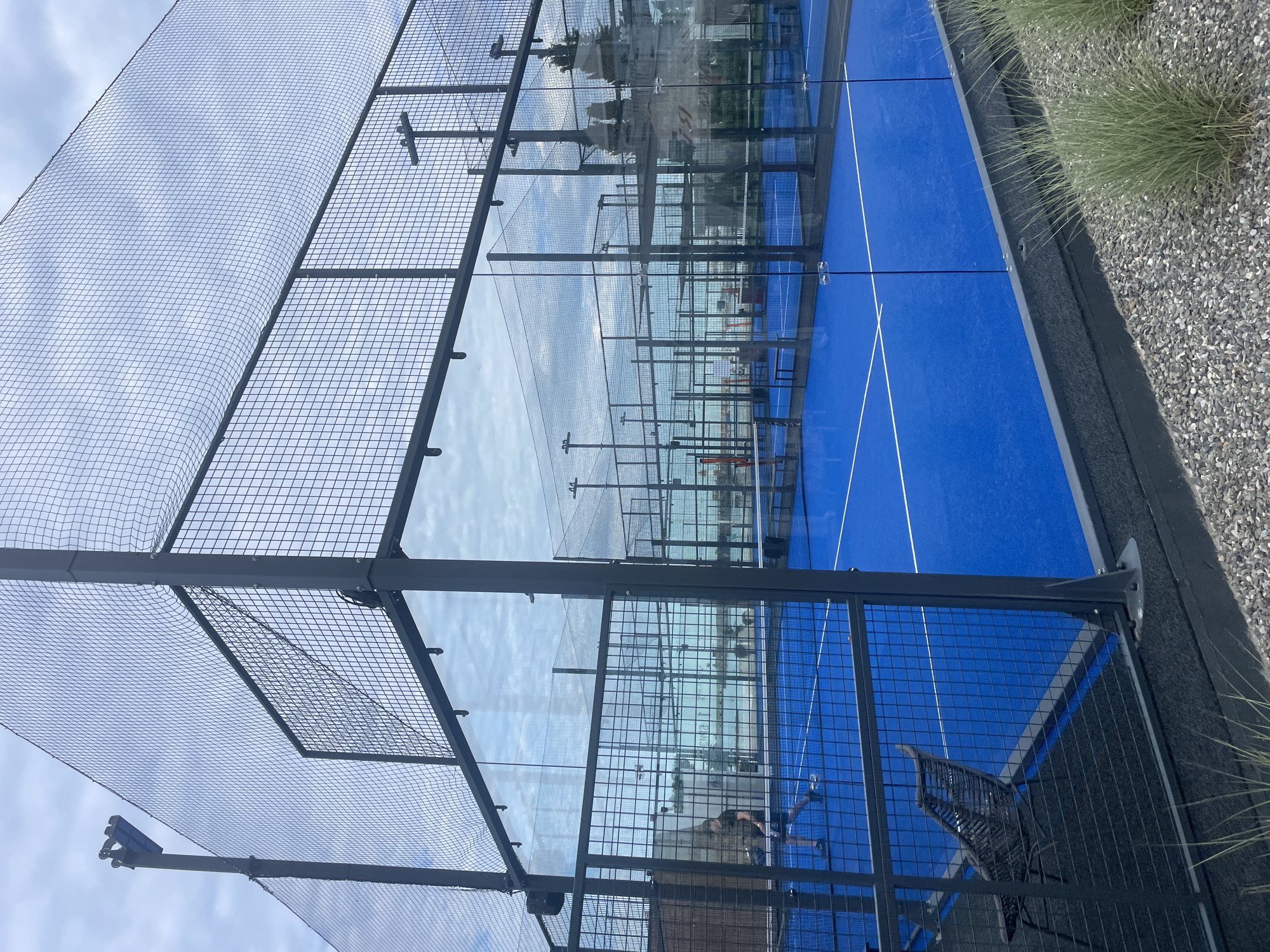 This is an eye-level, medium shot of an outdoor padel tennis court complex in Sochi, Russia, under a cloudy sky. The court is bright blue with white lines and surrounded by glass walls and metal fencing. In the foreground, a portion of a dark gray mesh fence is visible, with a black woven chair to the left. Beyond the fence, a blue padel court is laid out. In the middle ground, a young male wearing a black t-shirt and shorts is playing padel, holding a paddle. Other padel courts are visible in the background, along with more fencing, netting, and distant buildings and trees, suggesting an urban or resort setting. The overall scene is clean and modern, with a focus on the sports facility.