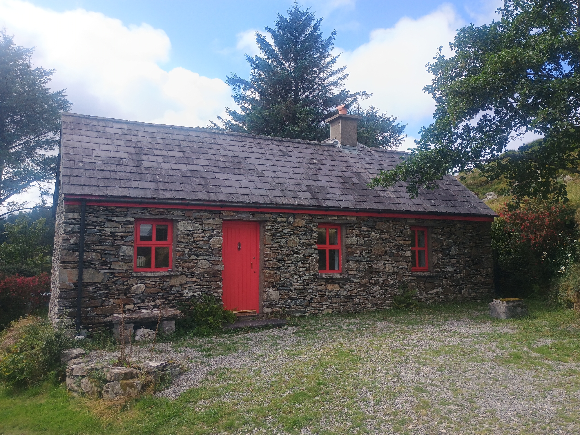 A rustic stone cottage with red window frames and a red door is pictured under a bright, partially cloudy sky. The cottage has a grey slate roof and a chimney made of brick. In front of the cottage is a gravel yard with patches of grass. To the left of the cottage, a stone bench sits outside, and to the right, a low concrete structure is visible. Trees and vegetation surround the cottage, with a large evergreen tree behind it and other foliage to the sides. The scene appears to be in a rural or natural setting, with no people or animals present.