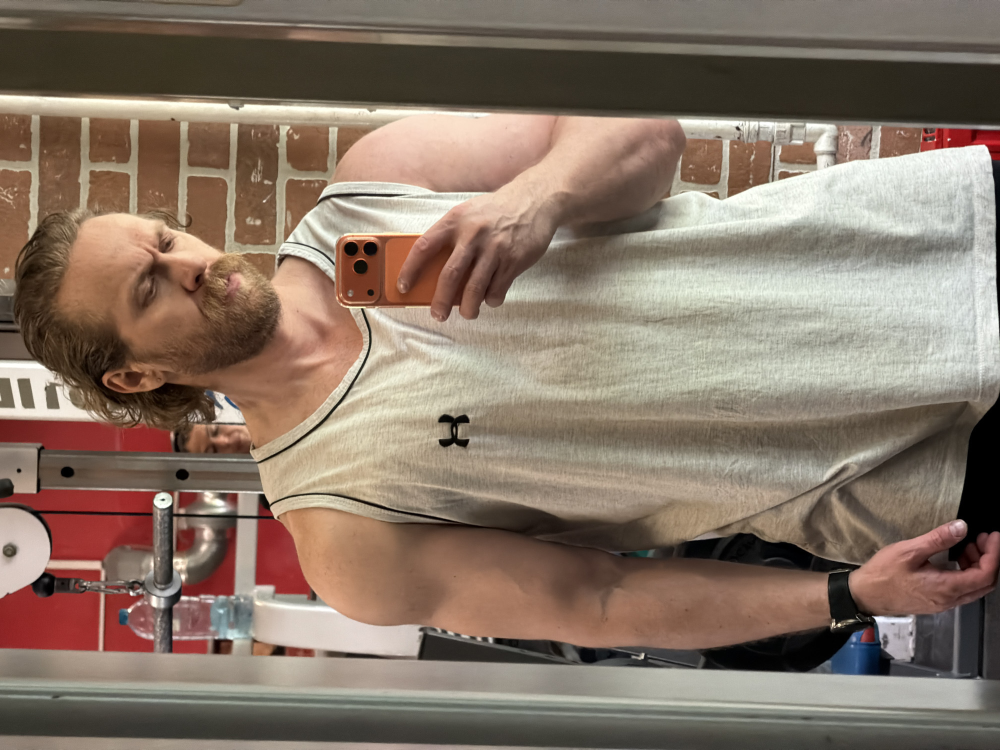 A man is taking a selfie in a mirror at a gym. He is wearing a light grey tank top with black trim and an Under Armour logo. He is holding a peach-colored smartphone. The man has fair skin, blonde hair, and a beard. In the background, there is a brick wall and exercise equipment. The gym appears to be indoors with fluorescent lighting. The image doesn't provide location information, so I cannot confirm if it is Saint Petersburg, Russia.