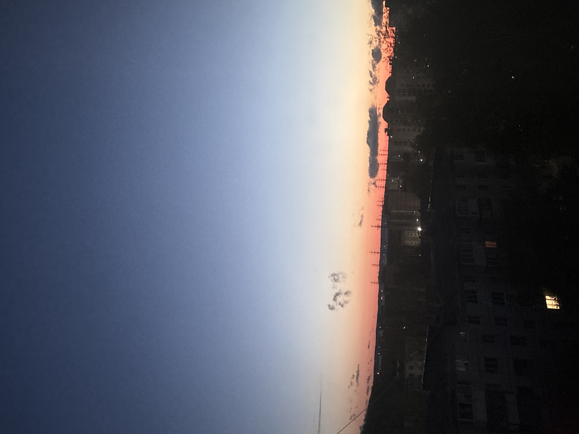 The image depicts a sunset over the city of Chaikovsky, Russia. The sky transitions from a deep blue on the left to a gradient of orange and pink hues near the horizon. The horizon is obscured by buildings, trees, and other urban structures. Silhouetted structures are seen along the skyline. Some clouds are visible, especially closer to the horizon. Streetlights and apartment windows are illuminated. The time of day is dusk.