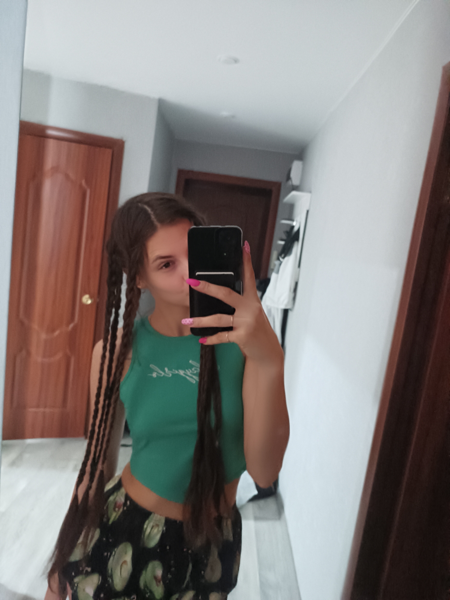 The image shows a person standing in an interior setting. The individual, presumably female, has long braided hair, a green tank top, and avocado-printed shorts. They are holding a smartphone in front of their face, likely taking a selfie in a mirror. The background reveals a hallway with two wooden doors and a white wall with shelves. The lighting appears to be artificial, coming from overhead sources. The location is described as being in the city of Unknown, Unknown.